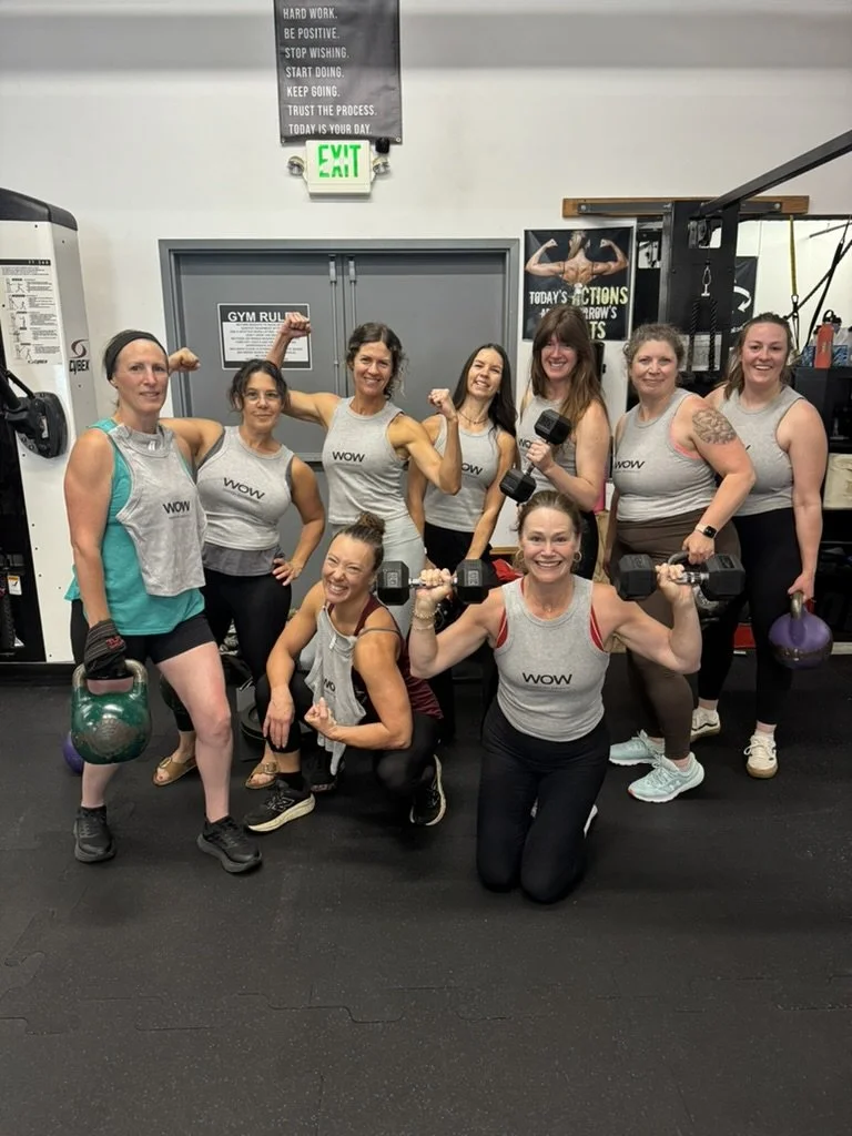 A woman in a black sleeveless shirt and black leggings is holding a dumbbell with both hands, performing a tricep extension exercise in a gym. Other women are visible in the background, also working out.