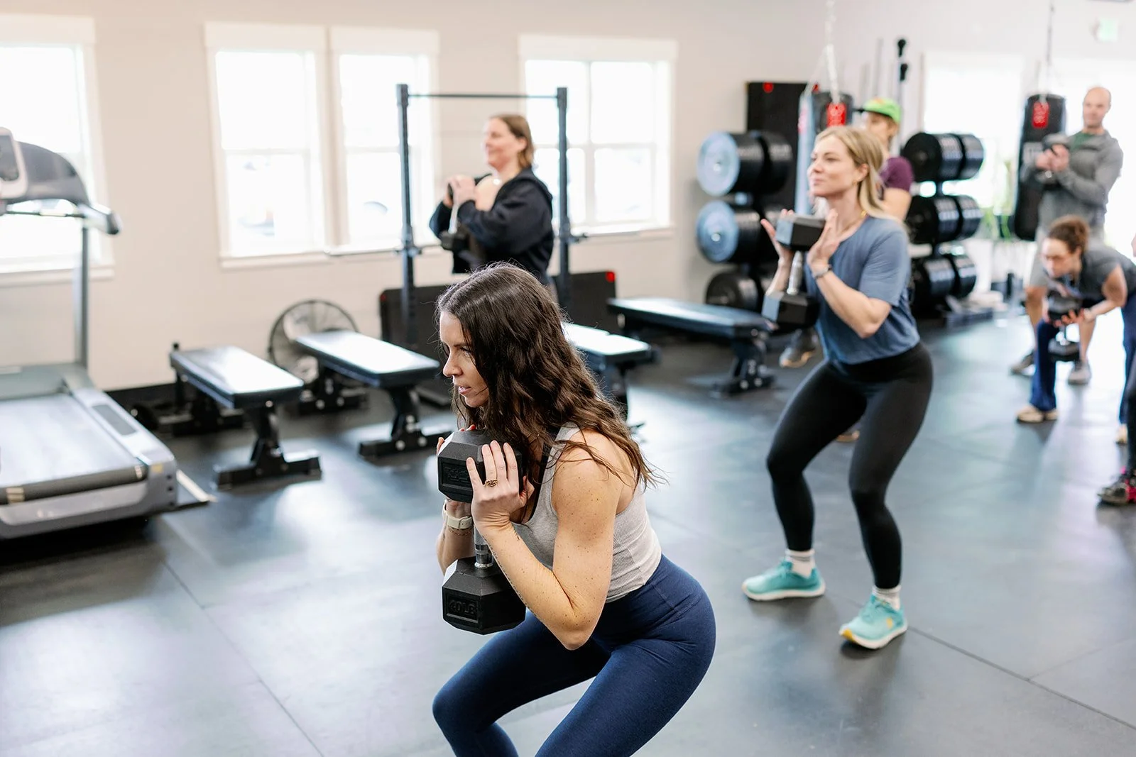 A woman in a black sleeveless shirt and black leggings is holding a dumbbell with both hands, performing a tricep extension exercise in a gym. Other women are visible in the background, also working out.