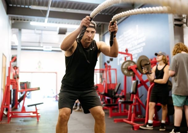 People working out in a gym, with one woman lifting weights and a trainer assisting, visible through a large mirror.