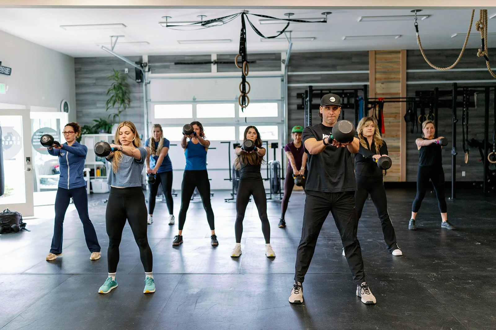 People working out in a gym, with one woman lifting weights and a trainer assisting, visible through a large mirror.