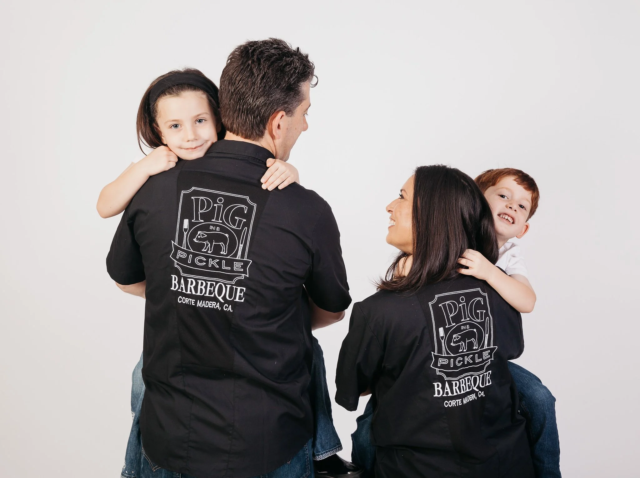 Pig in a Pickle family of four, including two young children, wearing matching black shirts with white logo and text, smiling and smiling and hugging each other against a plain white background.