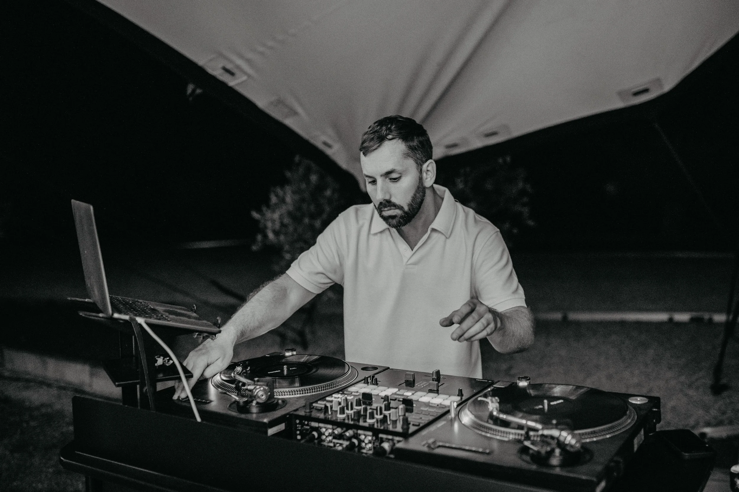 A man DJing at an outdoor event at night, operating a turntable and mixer under a canopy.