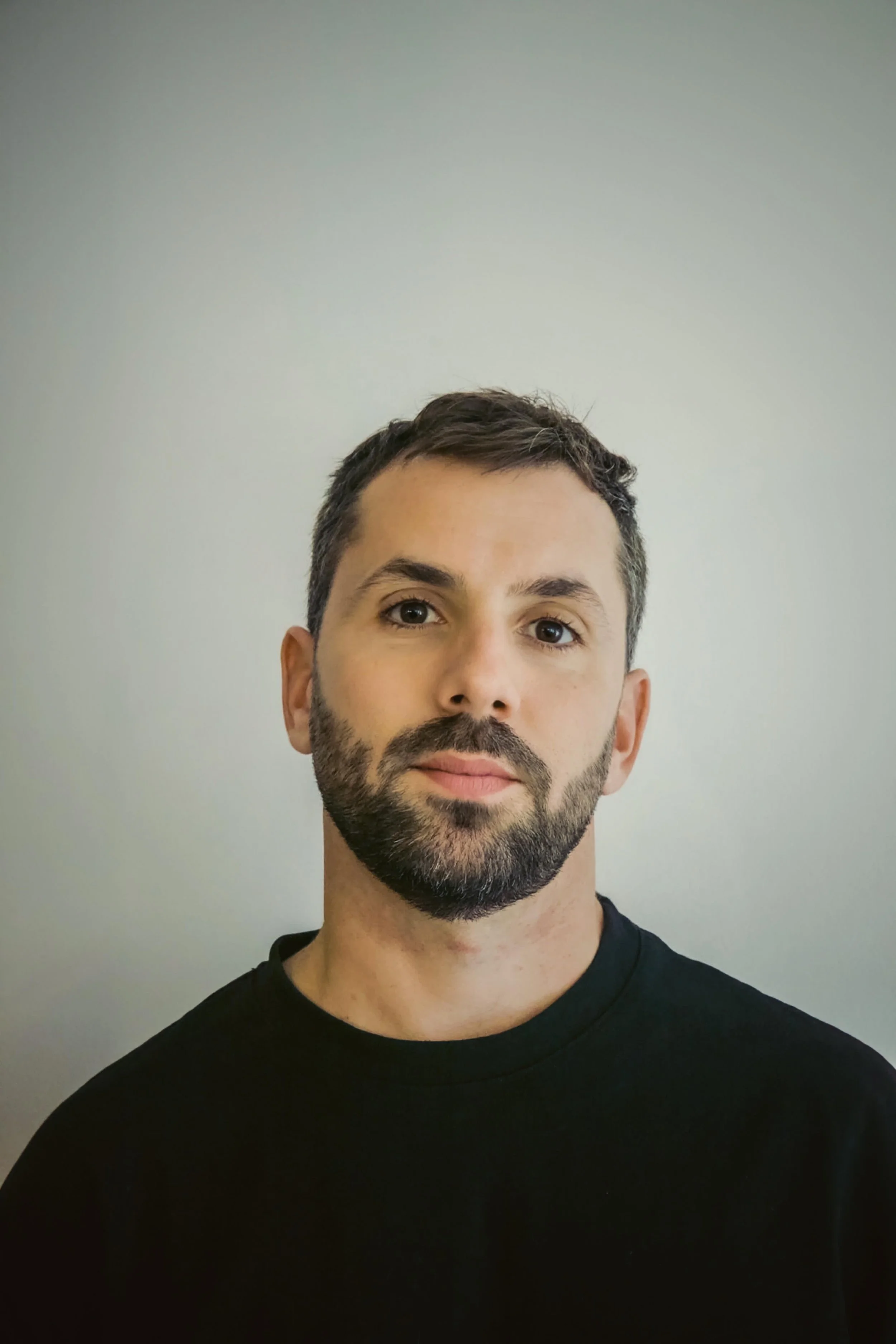 Portrait of a man with a beard and short dark hair, wearing a black shirt, standing against a plain light-colored background.