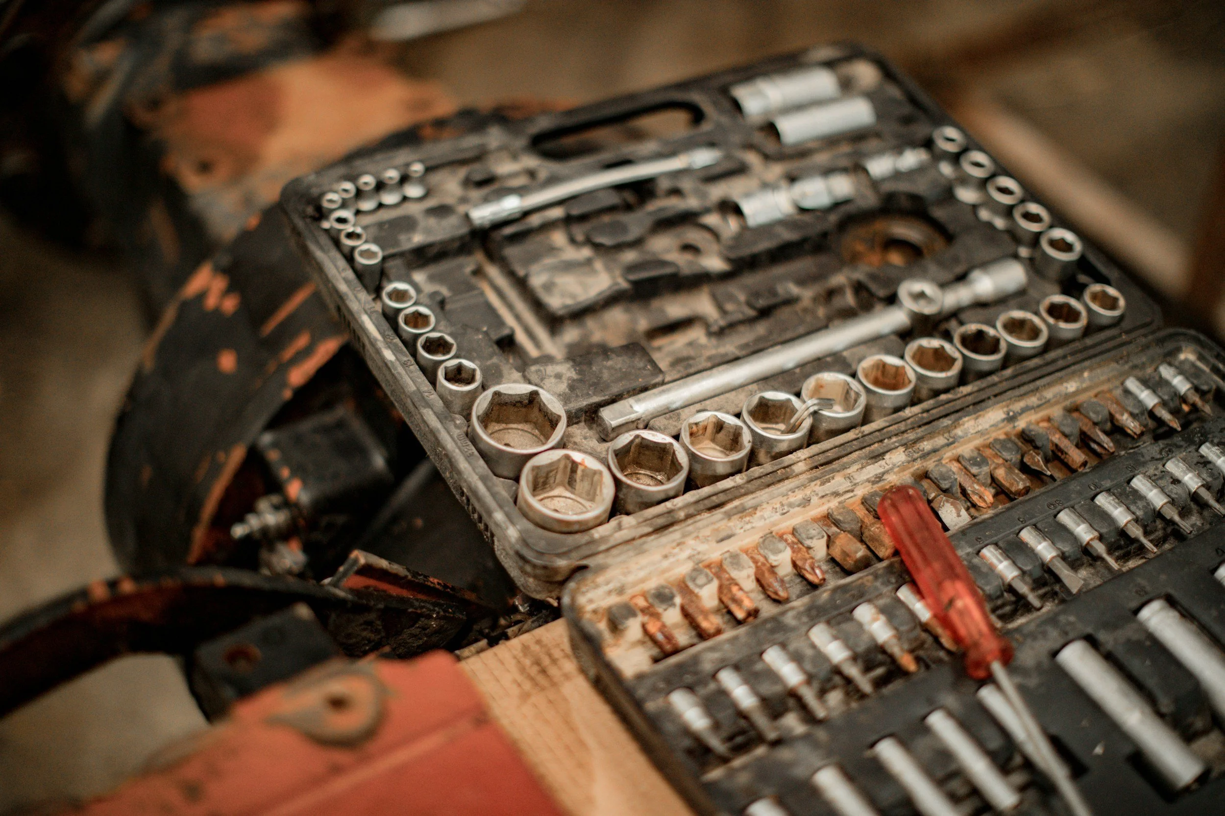 A toolbox with various sockets, ratchets, and screwdrivers, all covered in dirt and rust, sitting on a wooden surface.