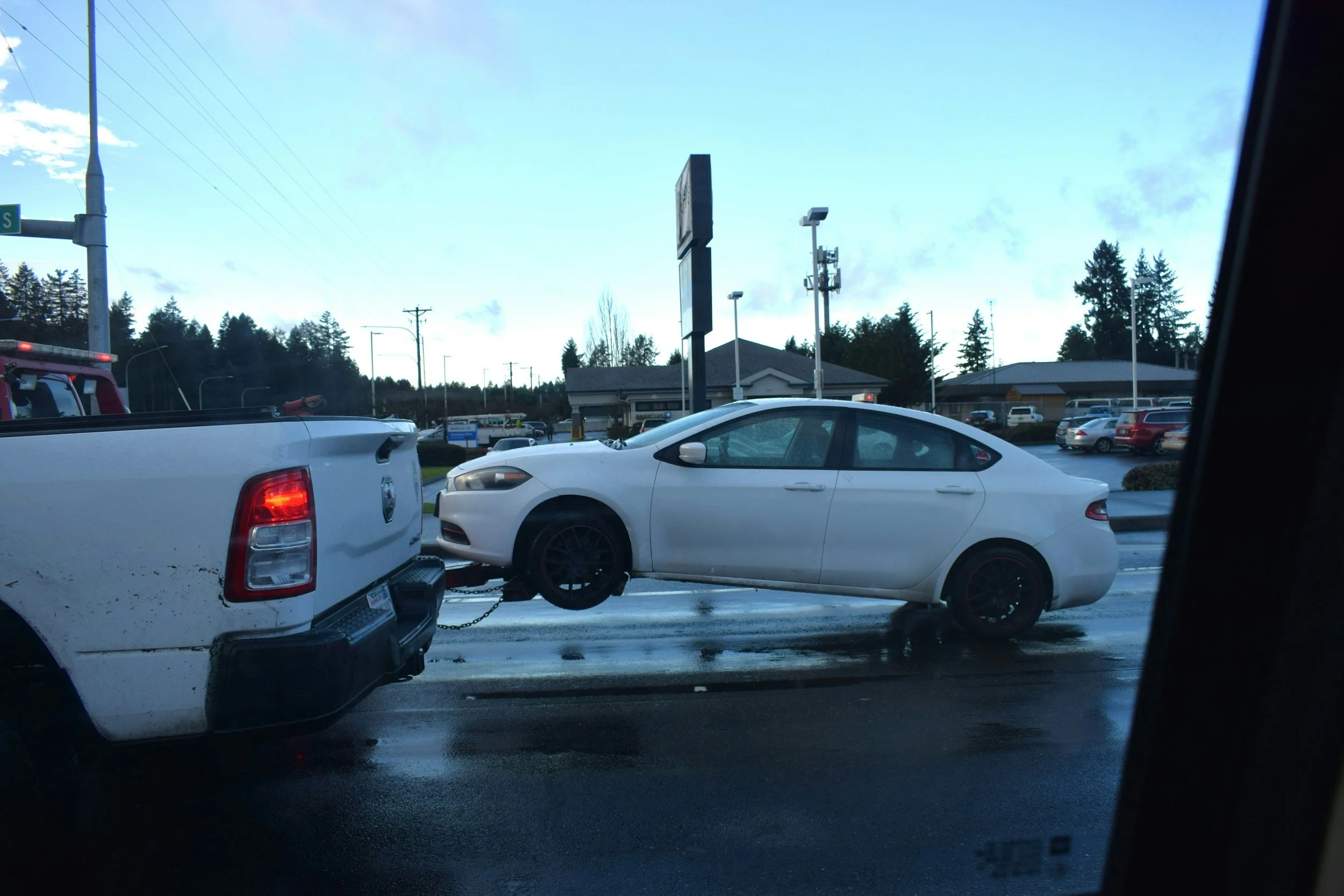 A white sedan is being towed by a white pickup truck on a wet road. The background shows a parking lot with cars and a building, and trees behind it. The sky is cloudy with some blue patches.