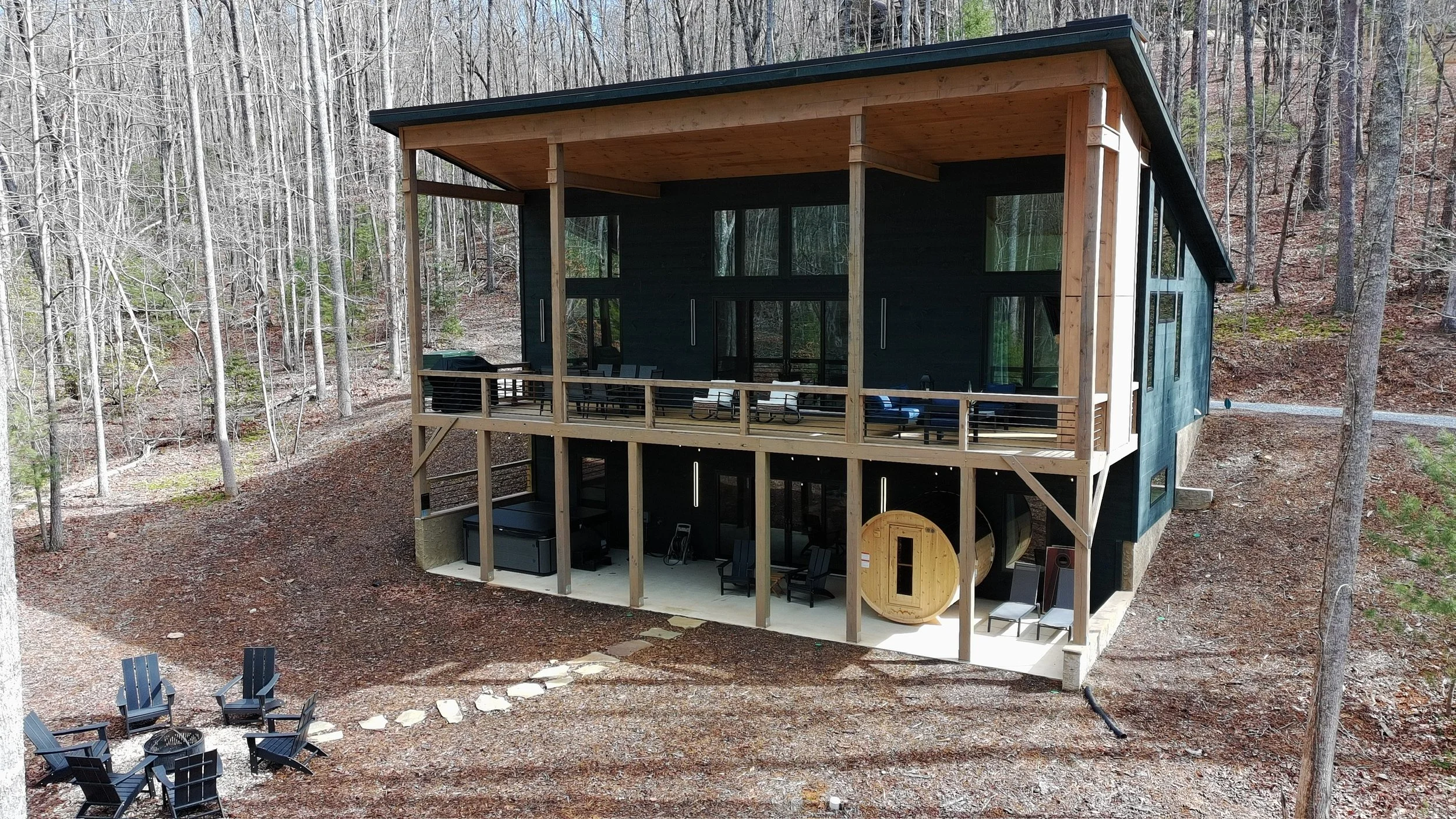 Modern multi-level cabin with large windows and a large spanning entertaining deck with grill and outdoor gas fireplace and barrel sauna, built into a hillside surrounded by trees