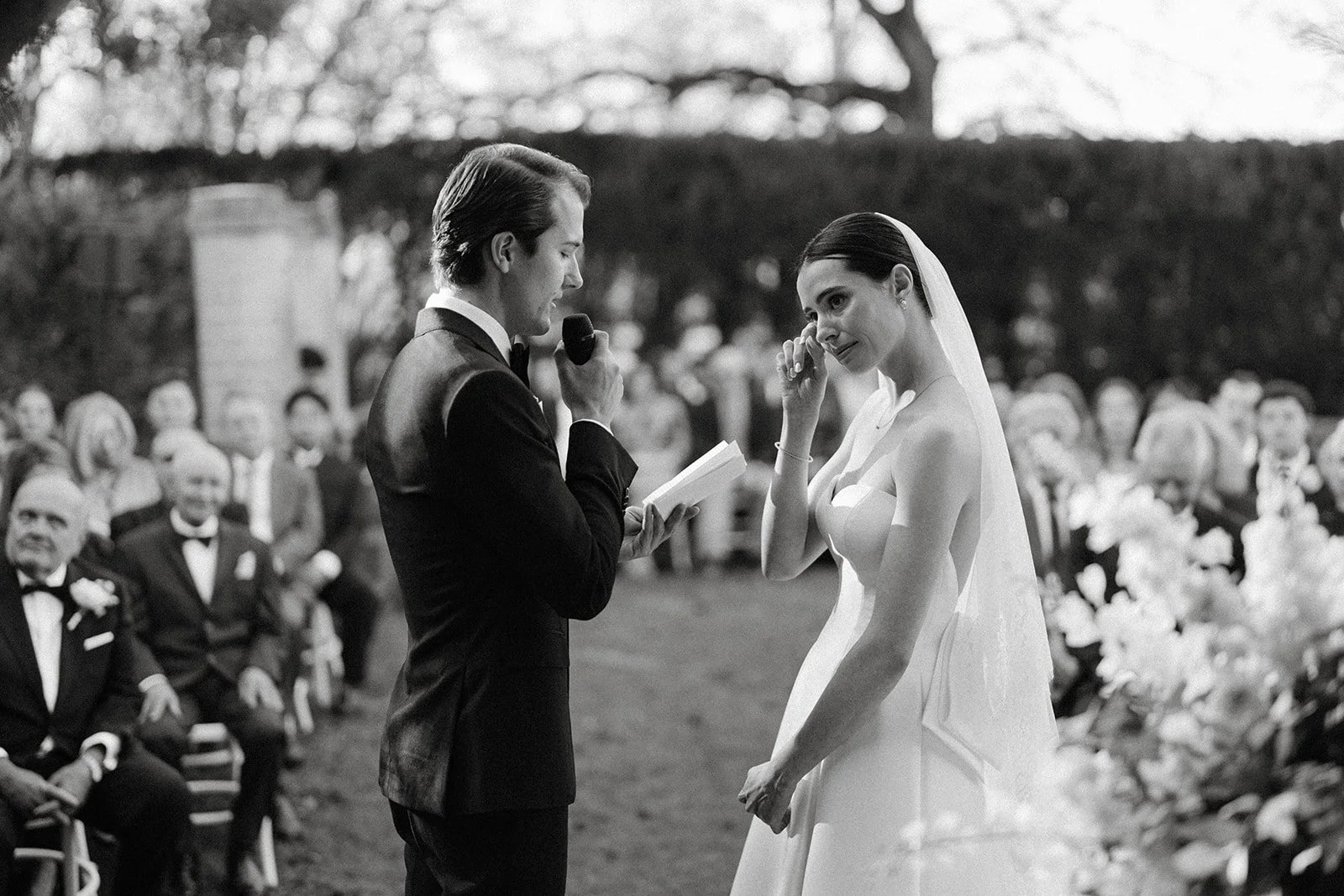A black and white photo of a wedding ceremony outdoors. A man in a tuxedo is reading from a book into a microphone, while a woman in a wedding dress and veil is wiping tears from her eyes. Guests are seated in the background.