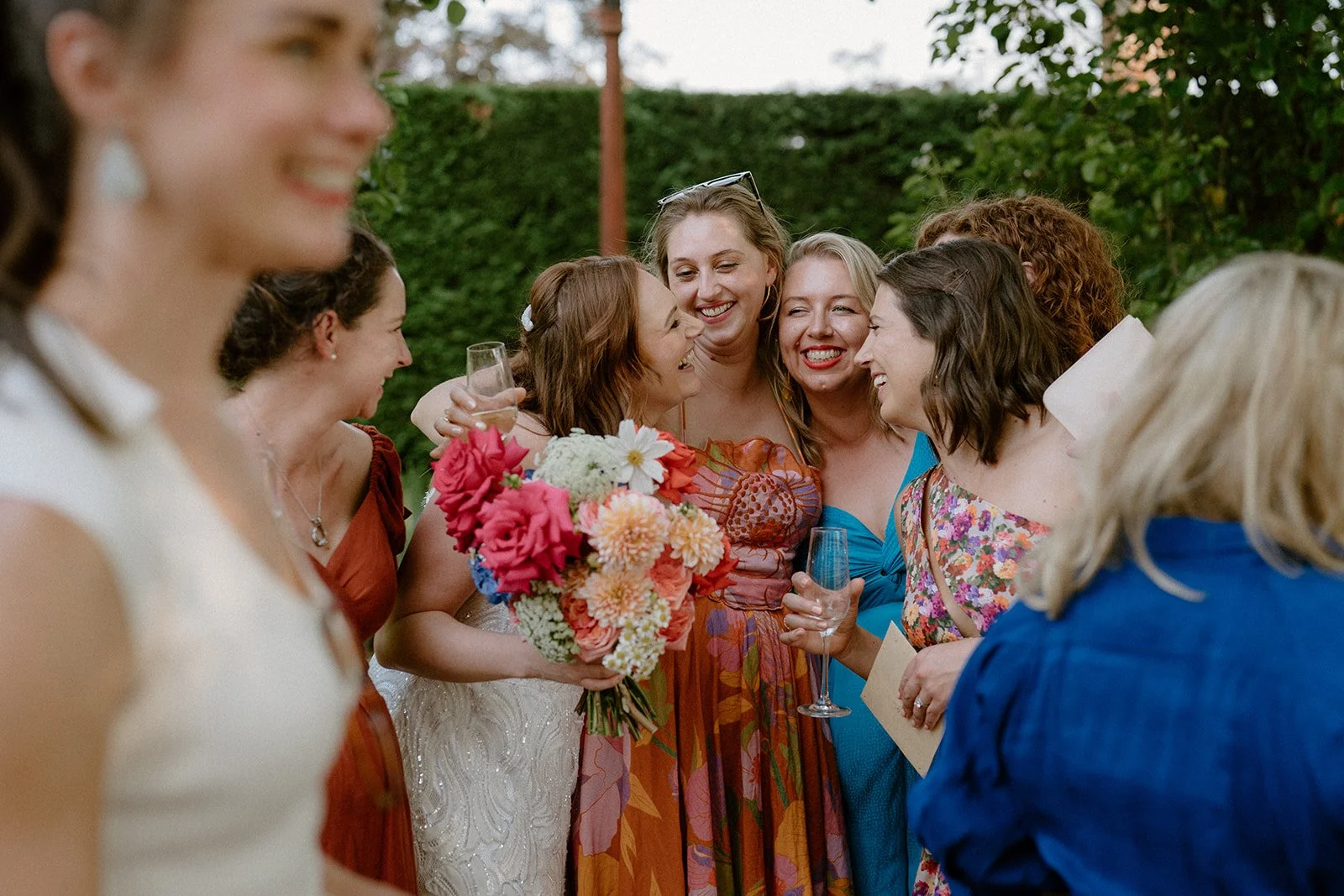 Group of women at a Homestead Berry wedding hugging and smiling, with one holding a bouquet of colourful flowers and some holding glasses of champagne.