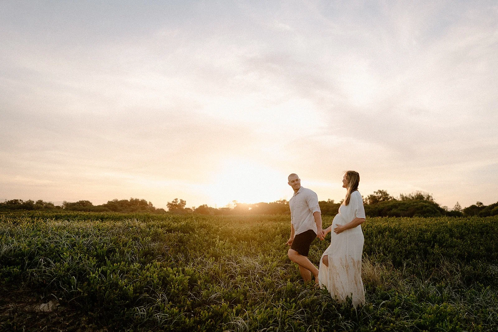 A couple during their maternity session, walking in long grass with a beautiful sunset in the background.