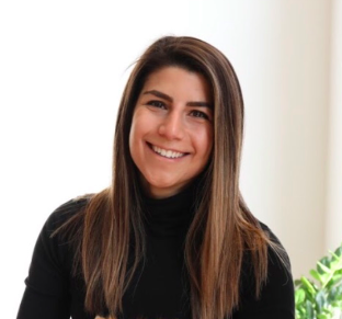 Person with long brown hair smiling, wearing a black turtleneck, indoor setting with a plant in the background.
