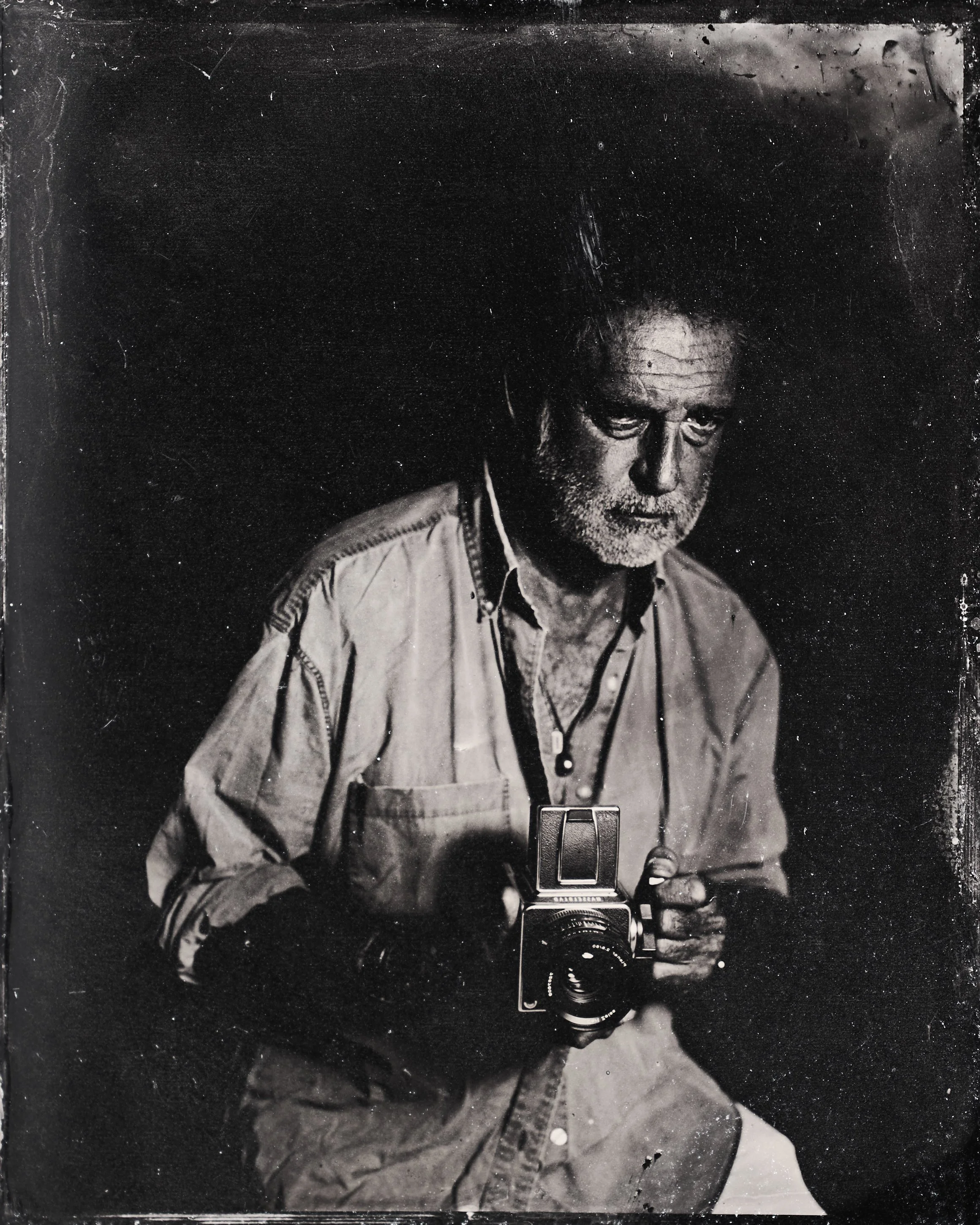 Black-and-white photo of a man with white hair and beard, wearing a button-up shirt, holding a vintage camera, looking down thoughtfully. Tintype Portrait photography Studio darkroom