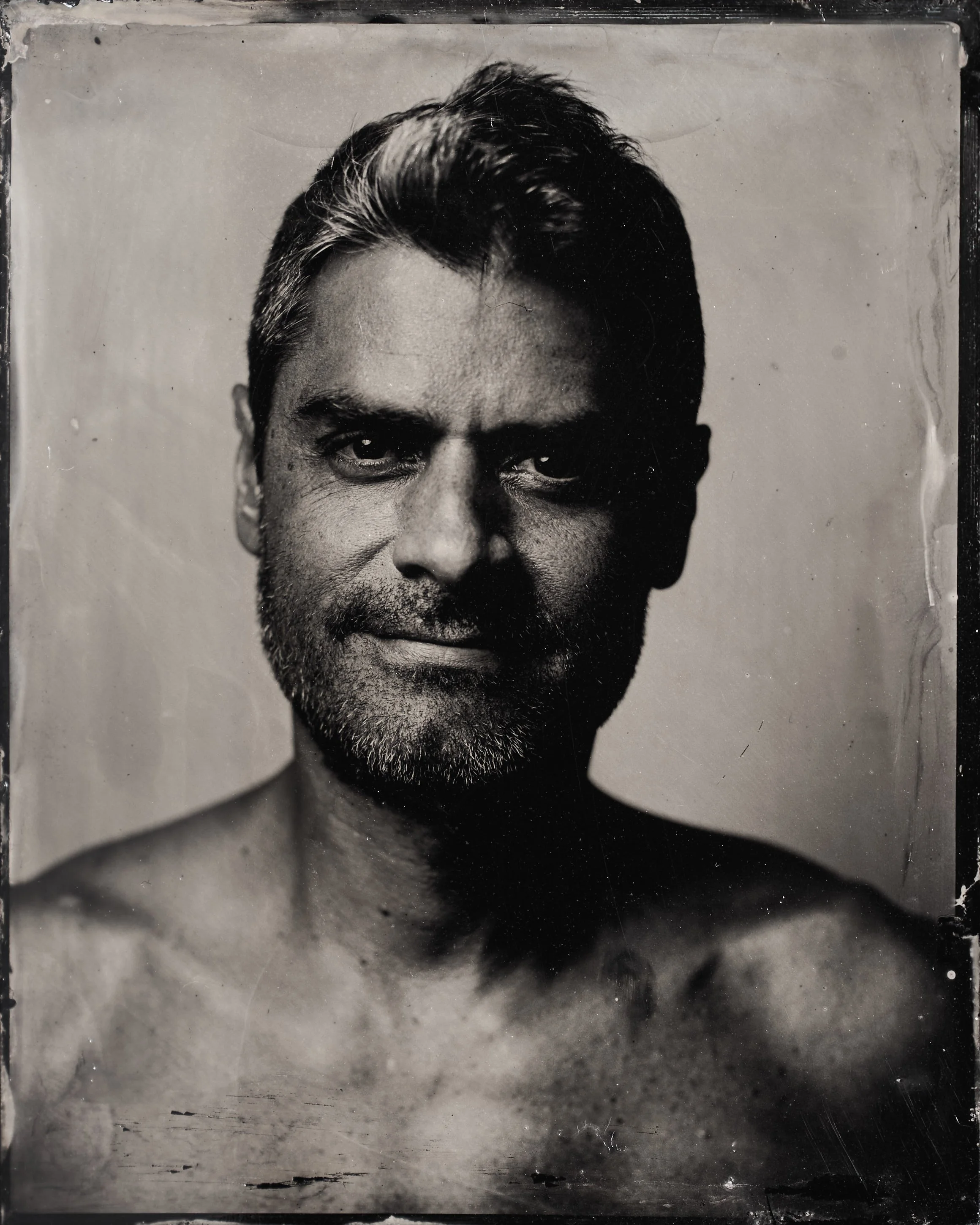 Black and white portrait of a shirtless man with a beard and styled hair, looking directly at the camera with a serious expression.