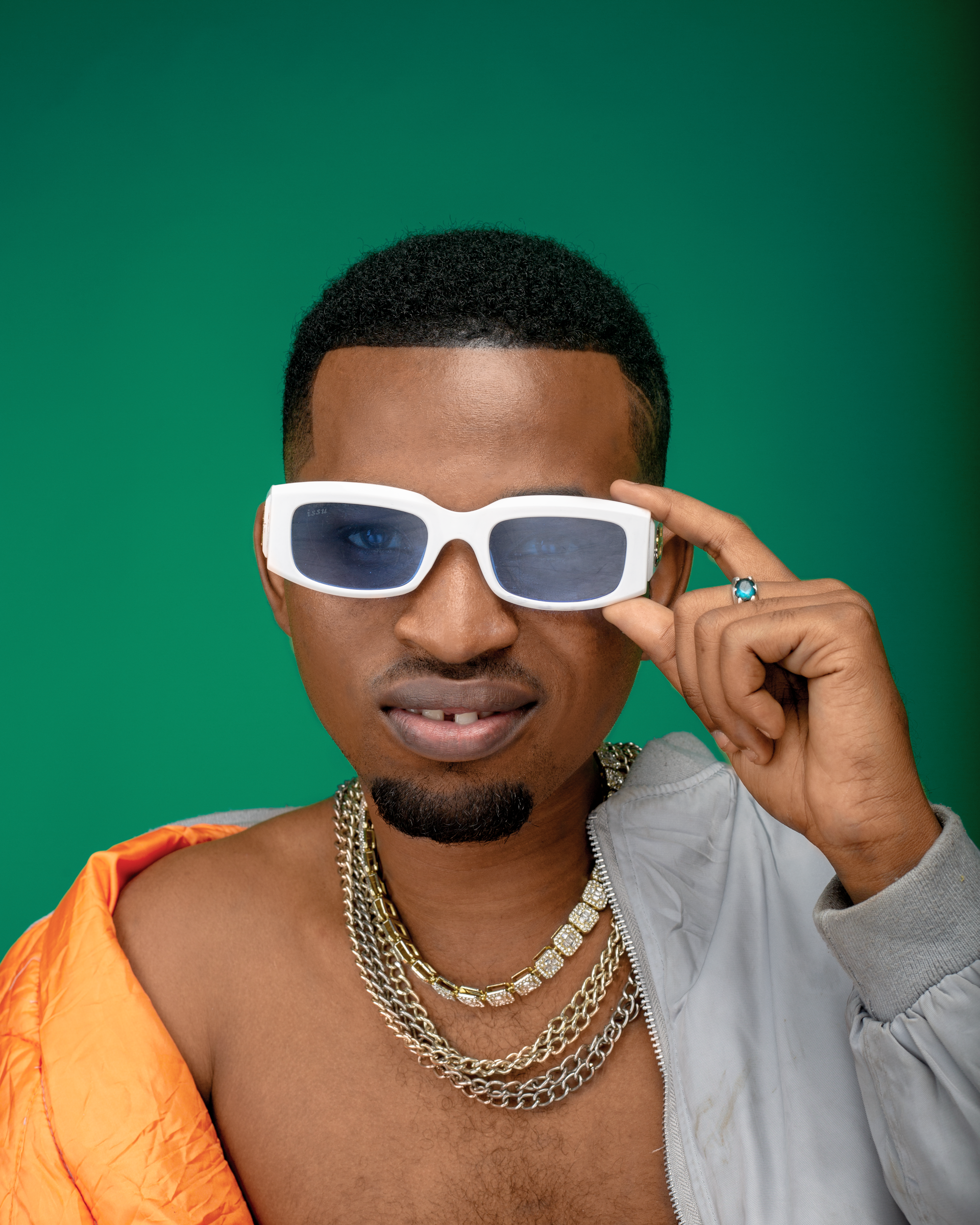 Person wearing white sunglasses, gold chains, and rings, with a green background.