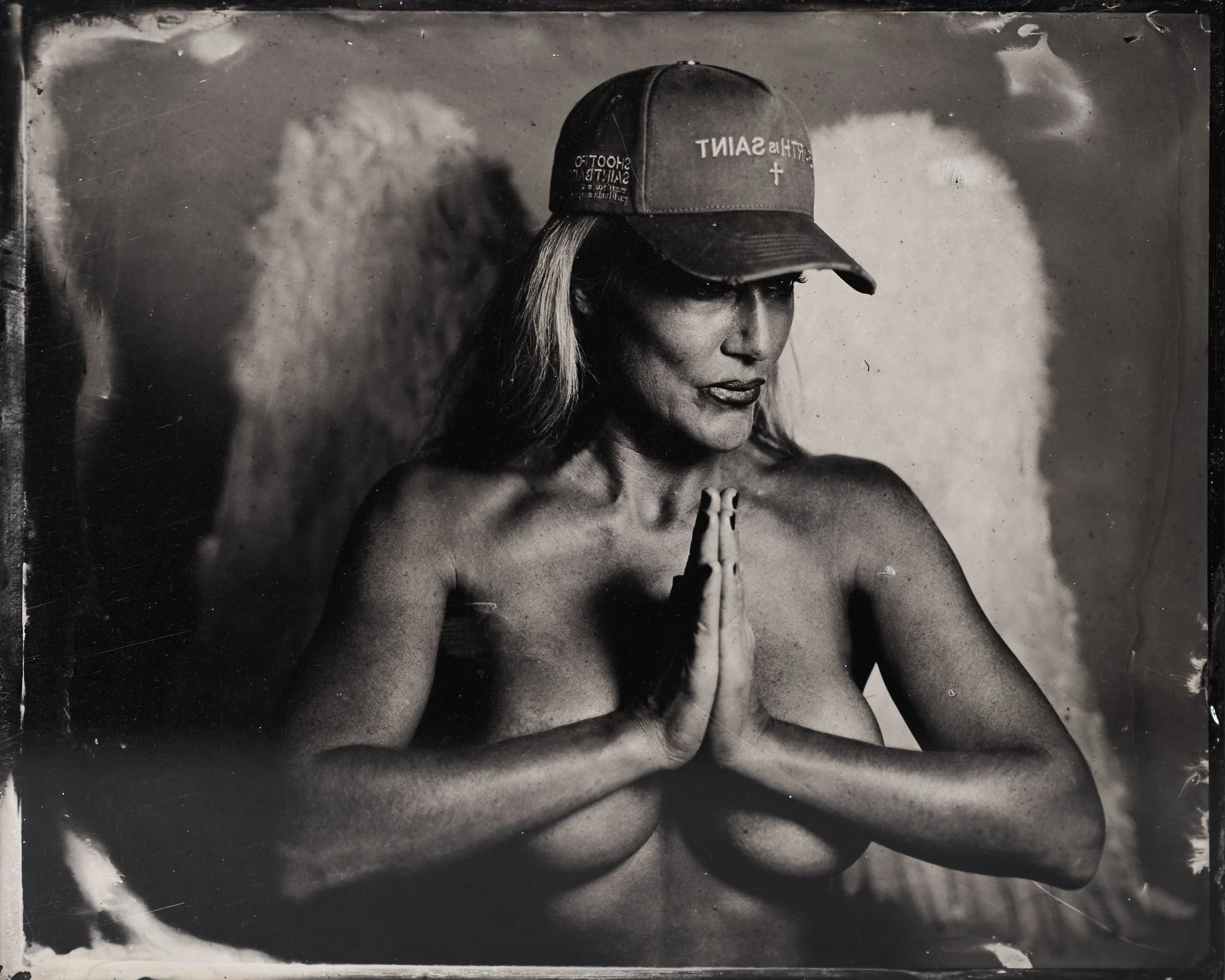 A topless woman with angel wings wearing a baseball cap, standing with hands pressed together in a prayer pose.