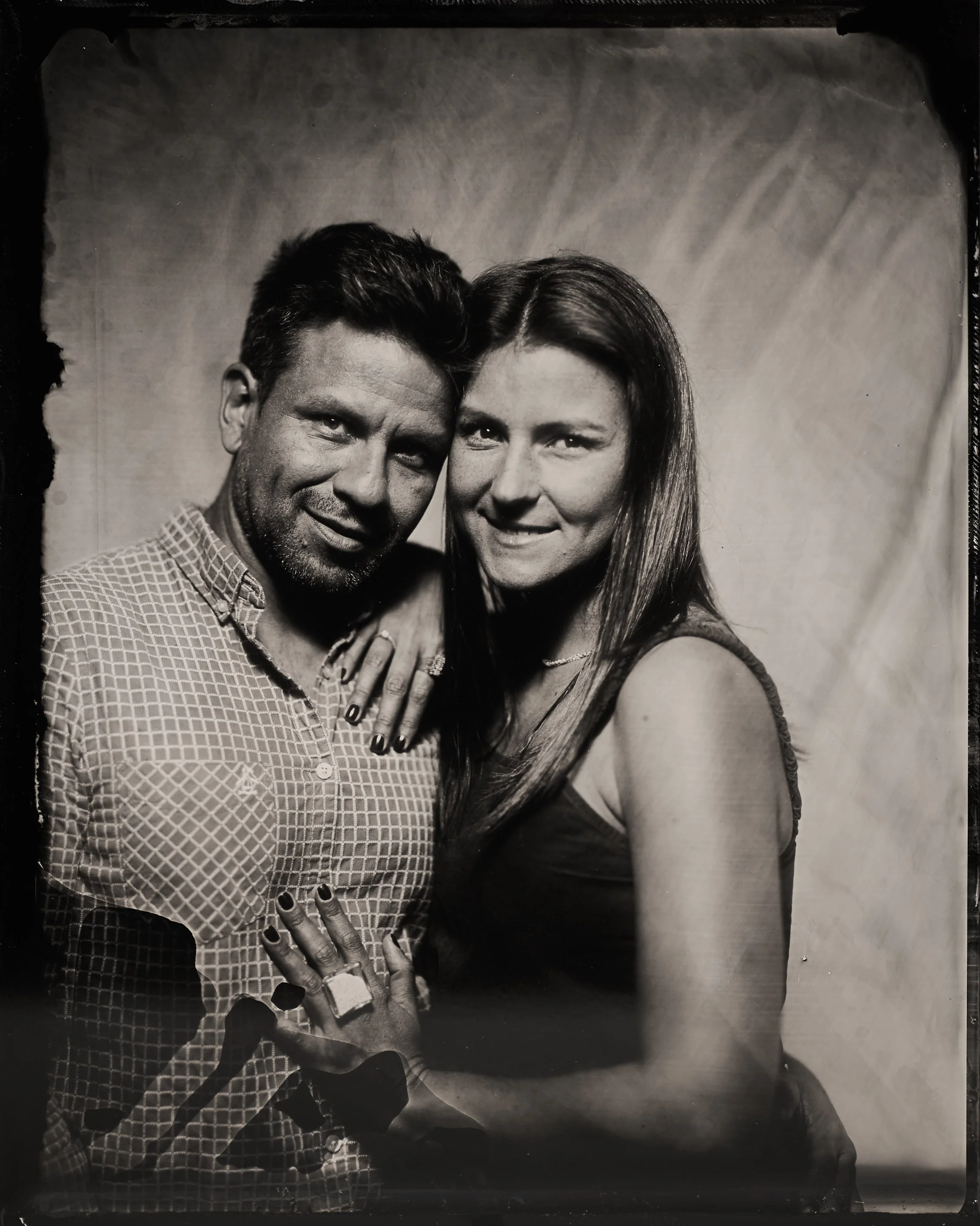 A black-and-white photo of a man and a woman smiling together, with the woman's hand resting on the man's chest. The woman has long hair and is wearing a sleeveless top, while the man has short hair and is wearing a patterned button-up shirt.