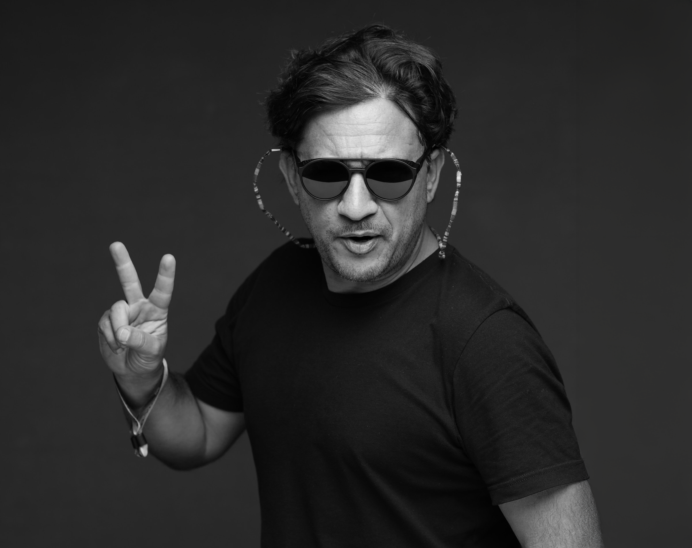 Black and white photo of a man wearing sunglasses, making a peace sign with his right hand, and looking at the camera.