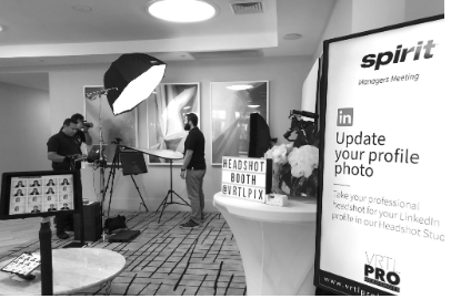 Photography setup at a corporate event with a ring light, camera, and a sign reading 'Headshot Booth' and 'VRTPIX'. A large screen displays a LinkedIn update alert for professional headshots.