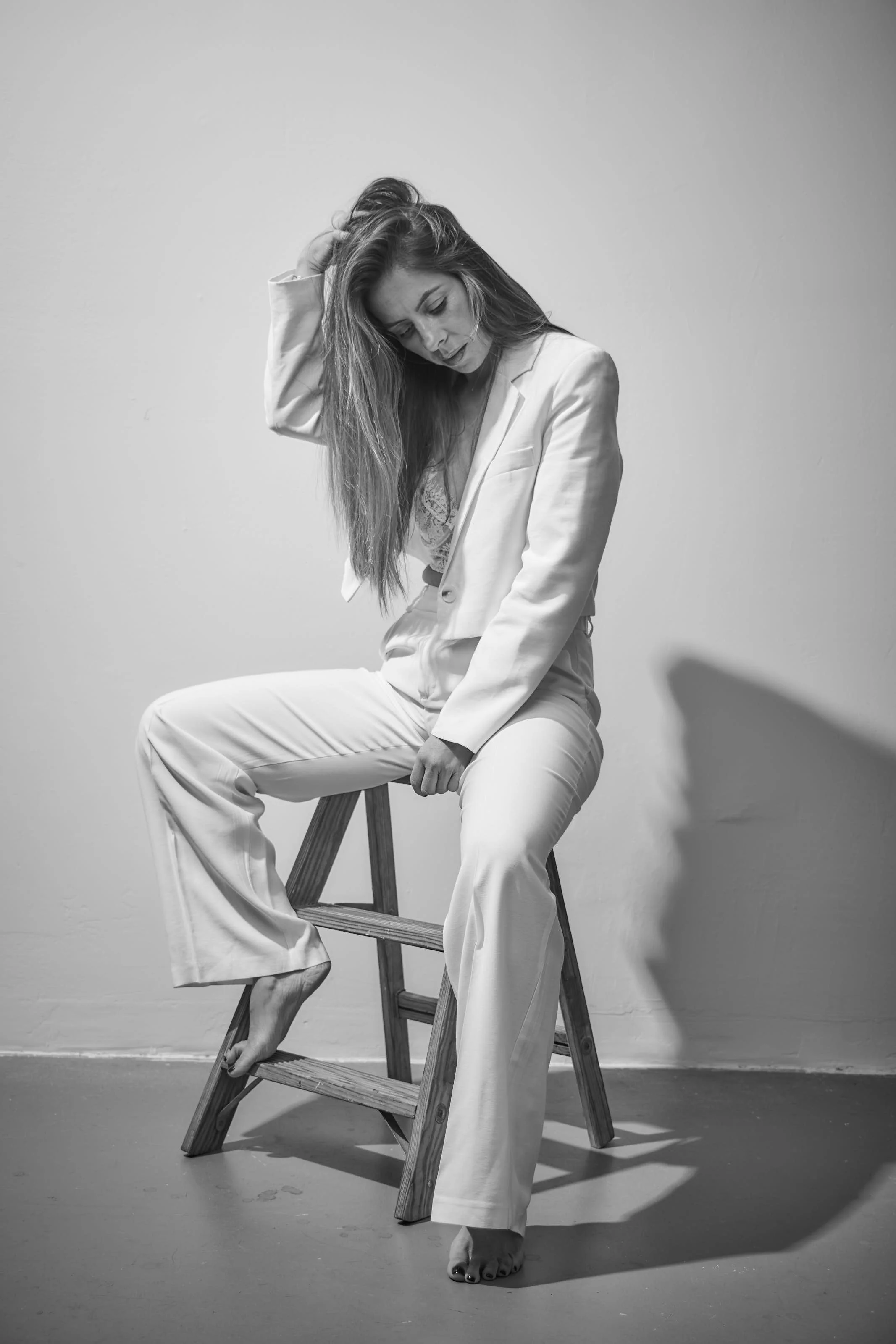 Black and white photo of a woman in a white suit sitting on a wooden step stool, with one foot on the ground and the other resting on the stool's lower step, her head tilted downward and her hand running through her long hair.