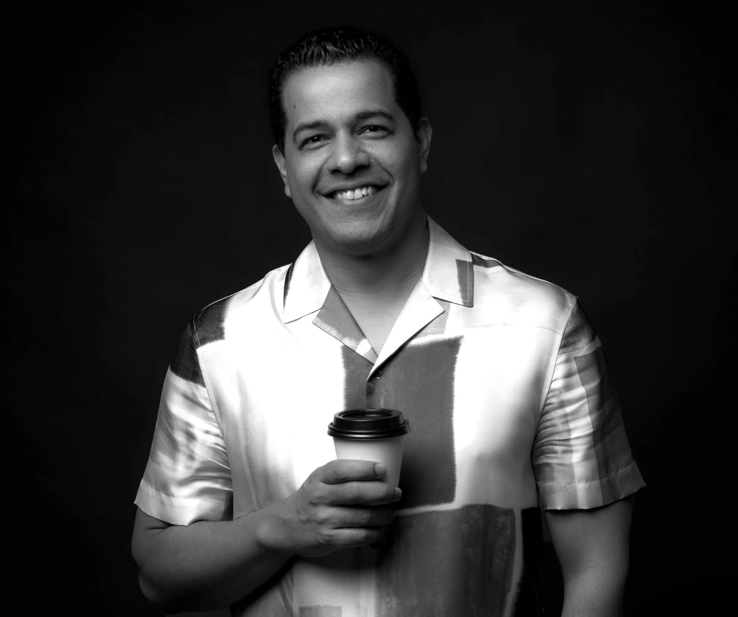 Black and white photo of a smiling man holding a coffee cup, wearing a short-sleeved, patterned button-up shirt, against a dark background.