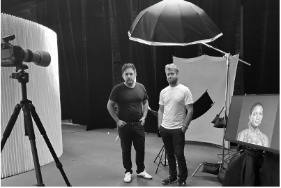 Two men standing in a photography studio with equipment, a large umbrella light, a backdrop, a camera on a tripod, and a framed portrait of a woman.