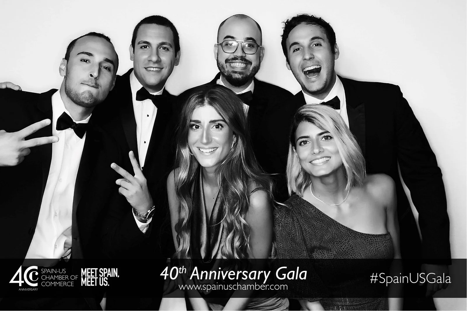 Group of six young adults dressed in formal attire, posing for a photo at a celebration event. Some are smiling, with one person making a peace sign. The image is in black and white.