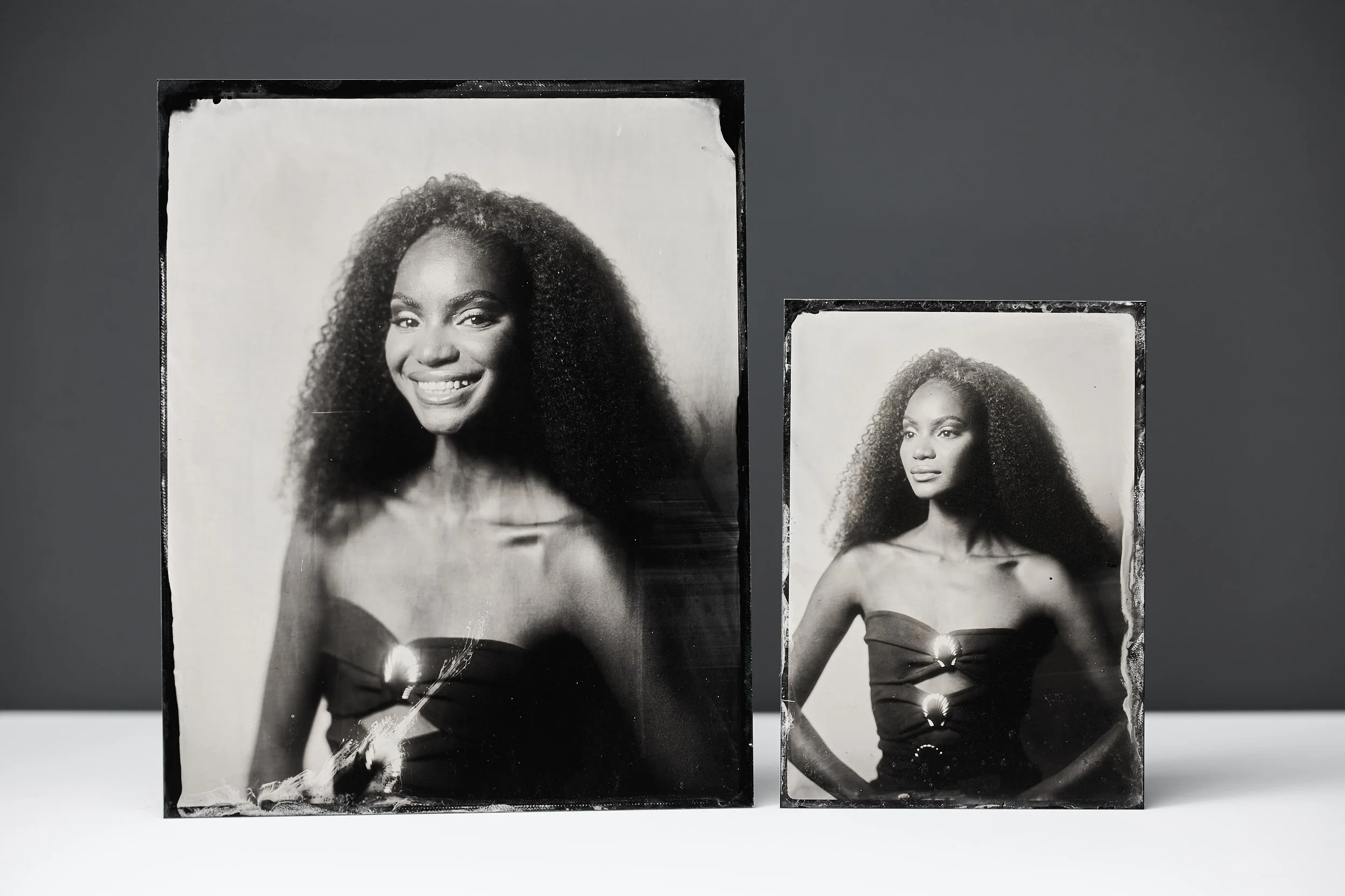 Two black and white photographs of an African American woman with curly hair, displayed on a white surface against a dark background. The larger photo shows her smiling with a strapless dress, the smaller photo shows her with a serious expression, we
