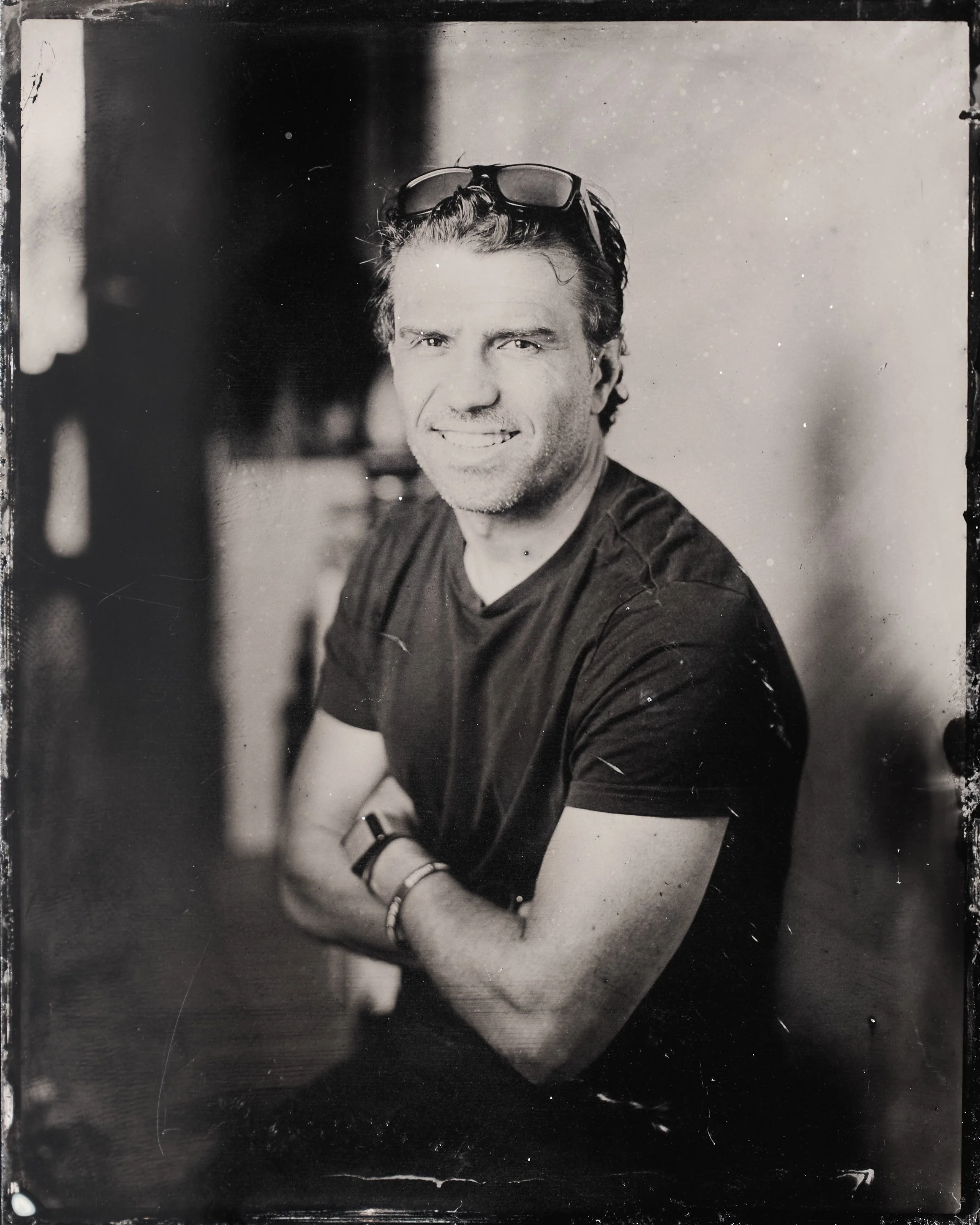 Black and white photograph of a smiling man with sunglasses on his head, wearing a dark t-shirt, sitting with arms crossed, in a casual setting.