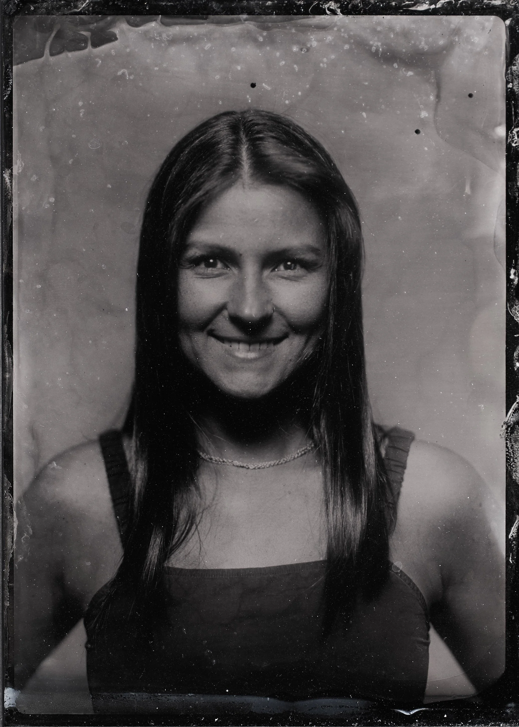 Black and white portrait of a woman with long hair, smiling, wearing a sleeveless top and a necklace. Tintype Portrait photography Studio darkroom