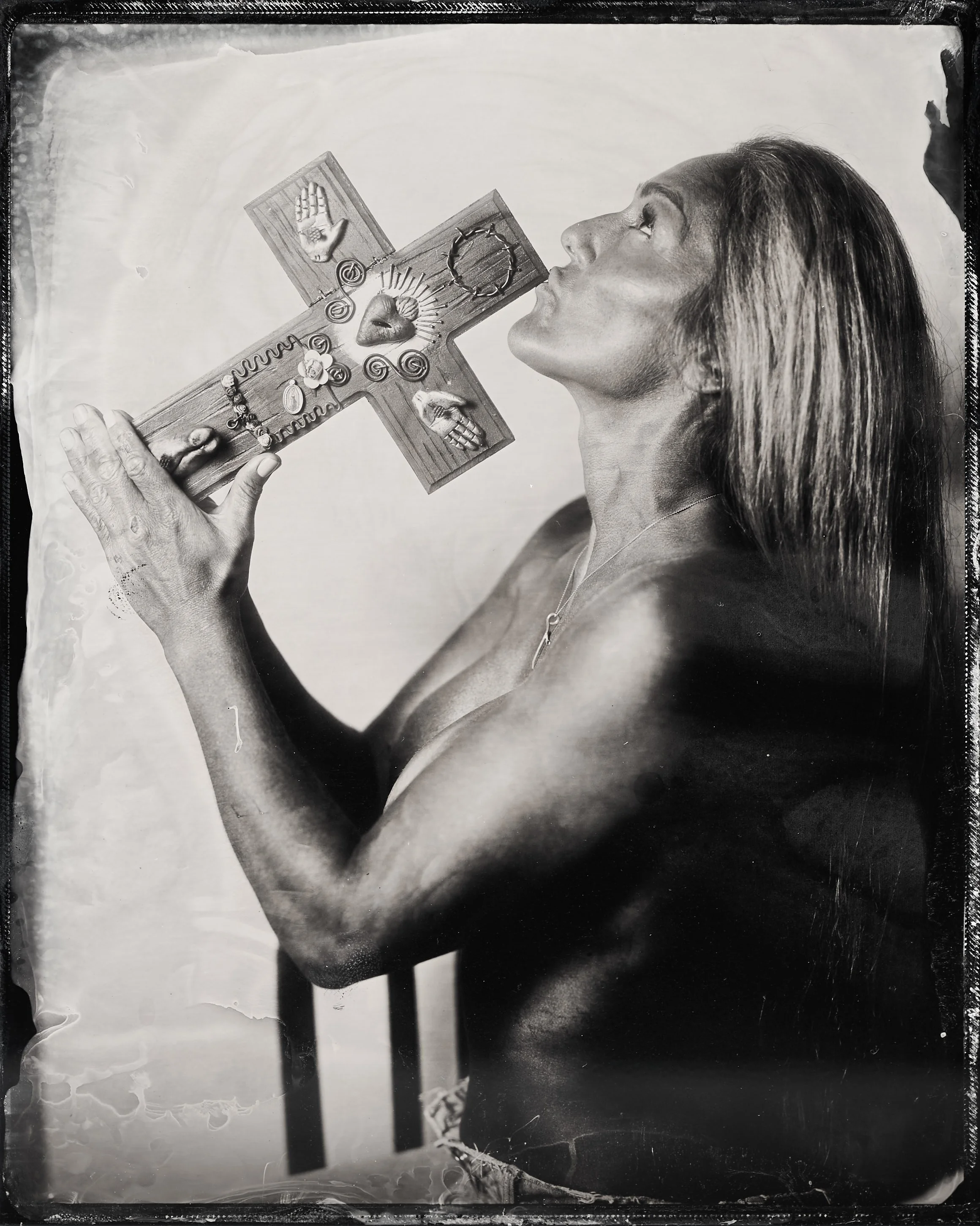 A woman with long hair holds a wooden cross decorated with various symbolic objects close to her lips, as if about to kiss it. Tintype Portrait photography Studio darkroom