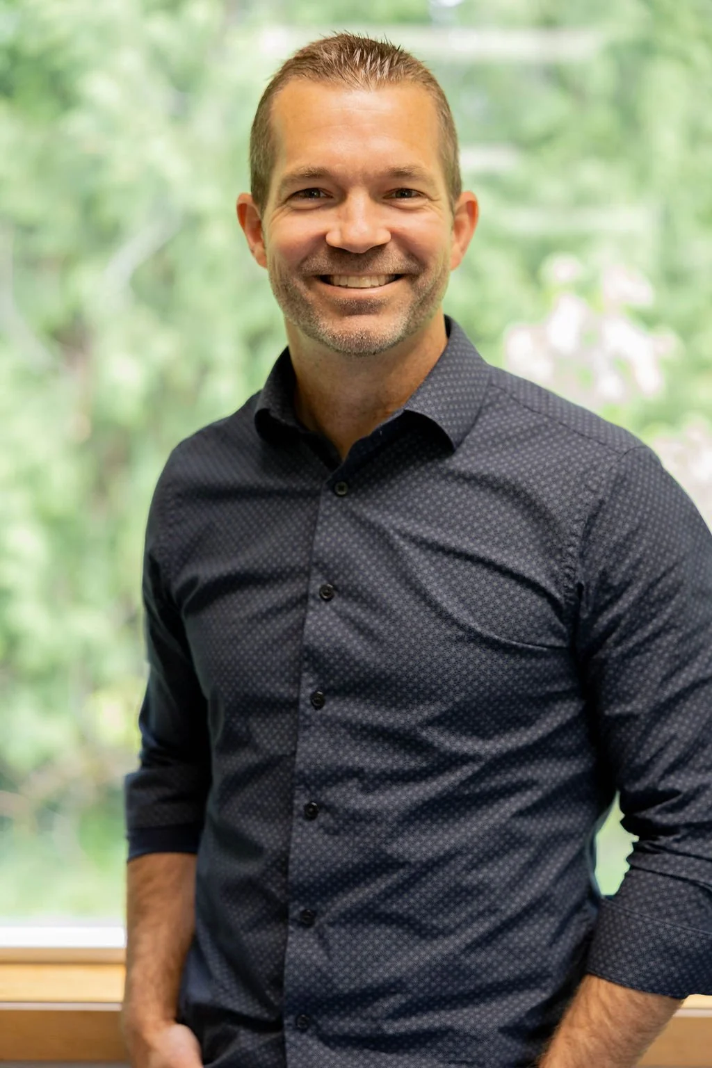 A man with short brown hair, a light beard, and a smile, standing in front of a window with green trees outside, wearing a dark patterned button-up shirt.