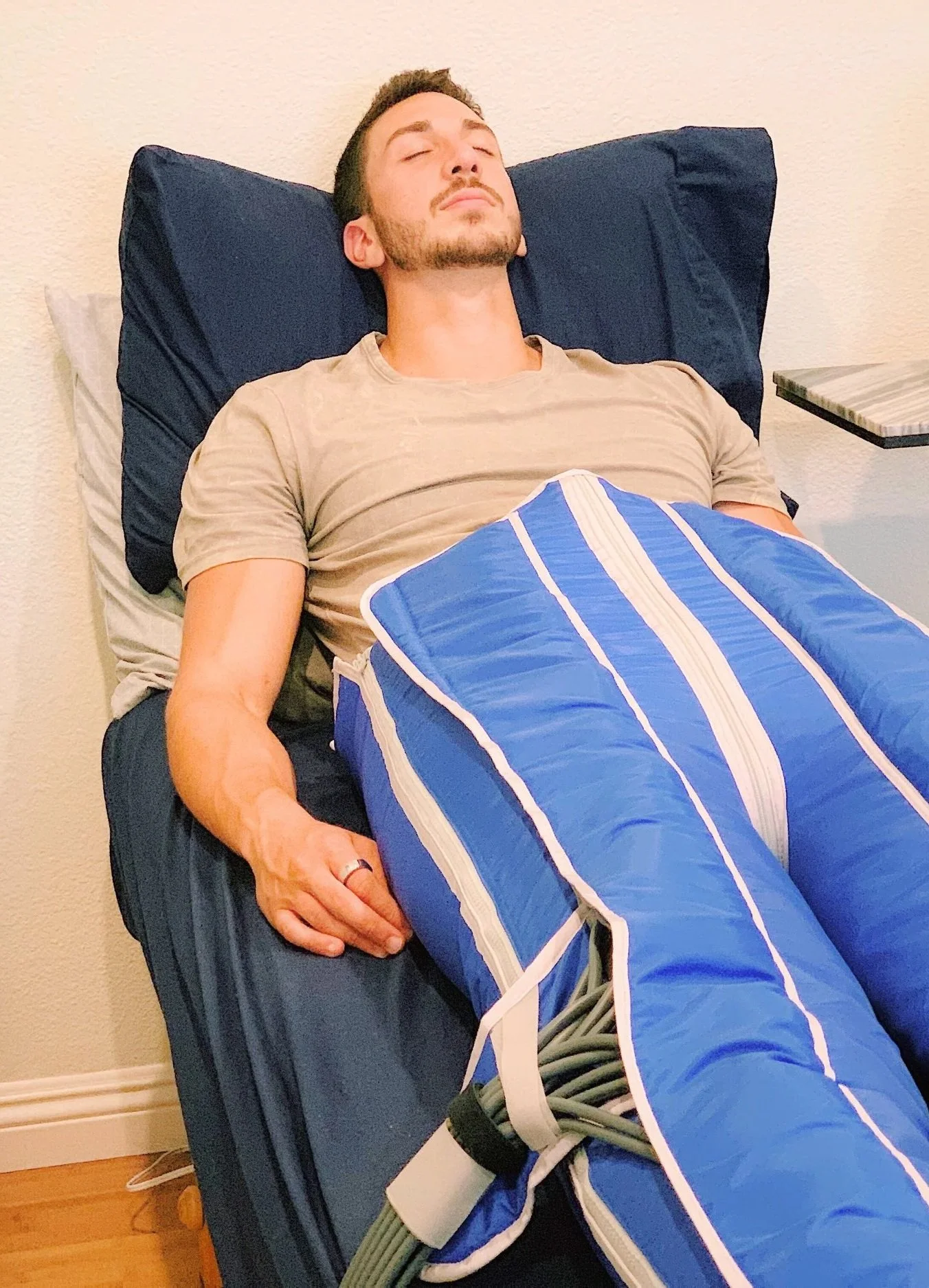 A man with a beard lies on his back while wearing medical-grade compression pants, receiving a sports massage treatment that helps with post-workout recovery. He wears a beige t-shirt and has his eyes closed. He seems relaxed.