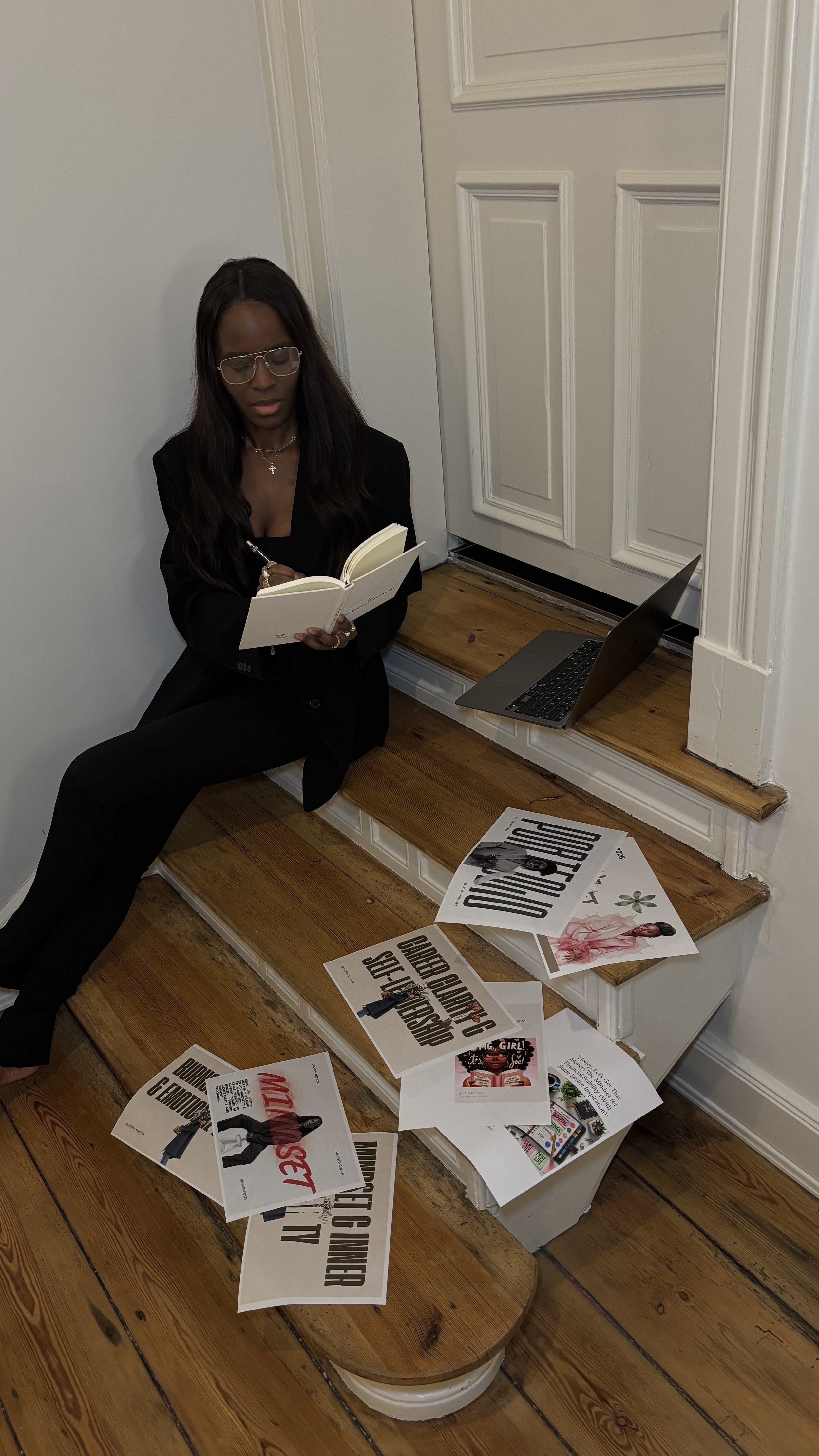 A woman with long dark hair, wearing glasses and a black blazer, sits on stairs reading a book. There are various printed papers and posters scattered on the stairs and a laptop placed on a higher step.