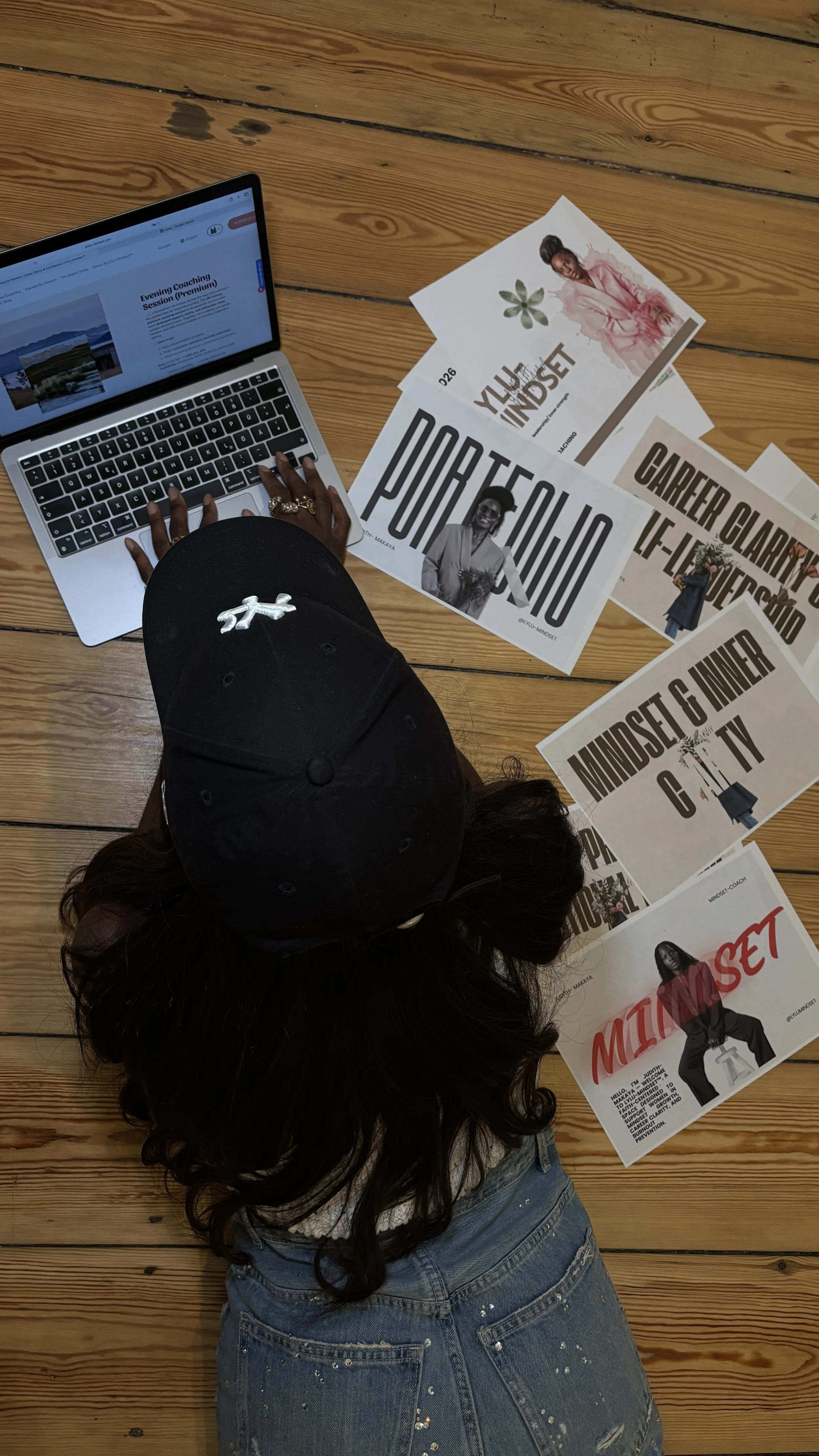 Overhead view of a woman with dark, curly hair wearing a black baseball cap and blue jeans, working on a laptop on wooden floor surrounded by printed posters.