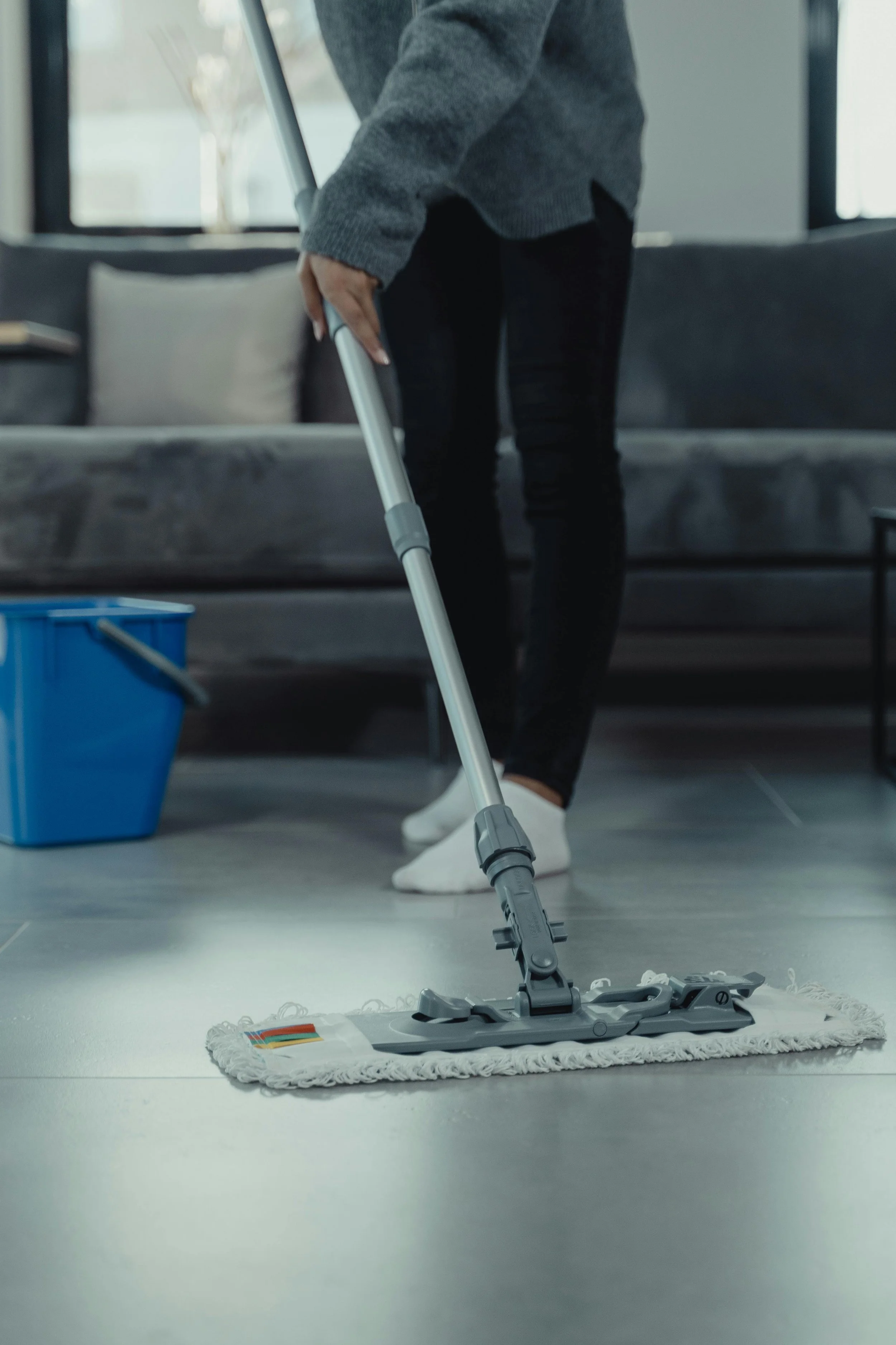 Person mopping a gray floor with a mop, wearing a gray sweater, black pants, and white shoes inside a home or office, with a gray sofa and a blue cleaning bucket in the background.