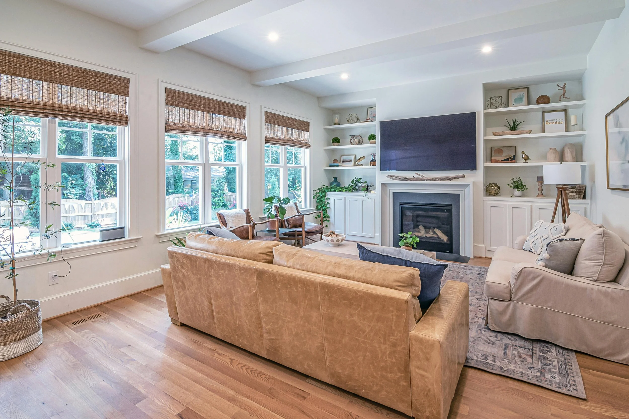 Bright living room with beige sofas, wooden chairs, a fireplace, and white built-in shelves, overlooking a backyard with tall trees and a wooden fence.