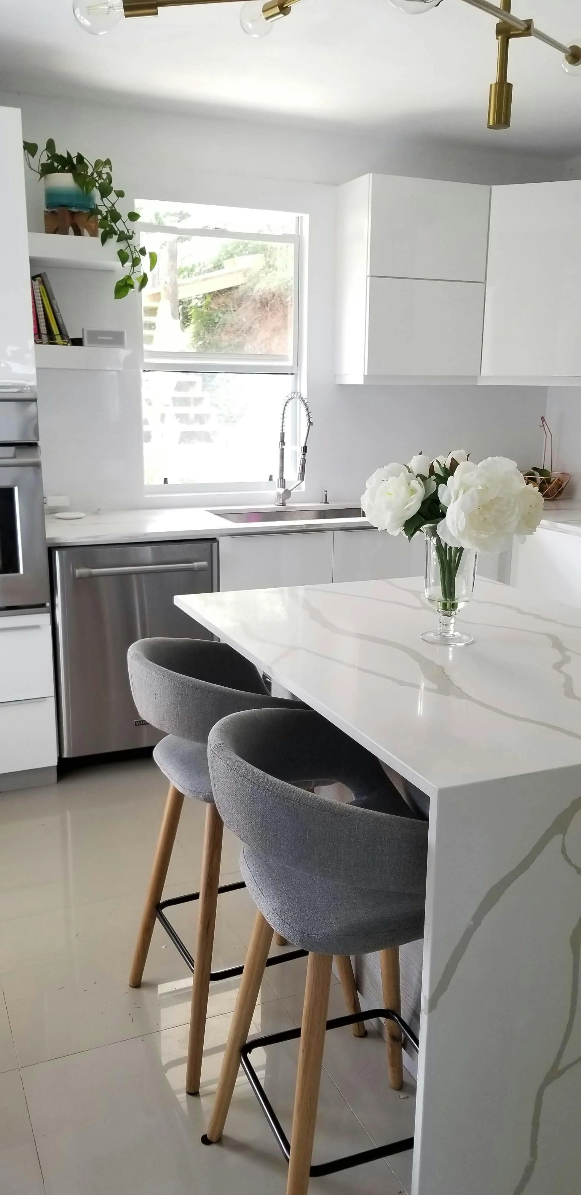 Modern kitchen with white cabinets, a window above the sink, stainless steel appliances, a white marble countertop, a glass vase with white flowers on the island, and two gray upholstered barstools with wooden legs.