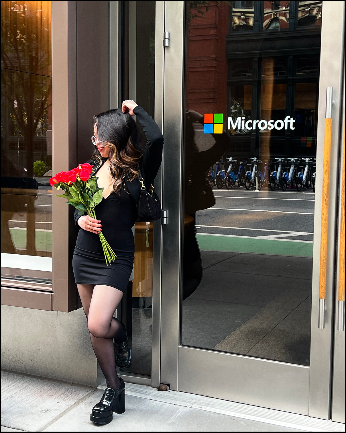 A woman in a black dress, black platform shoes, and glasses stands outside a Microsoft store, holding a bouquet of red roses, with a black purse over her shoulder, leaning against the door frame.
