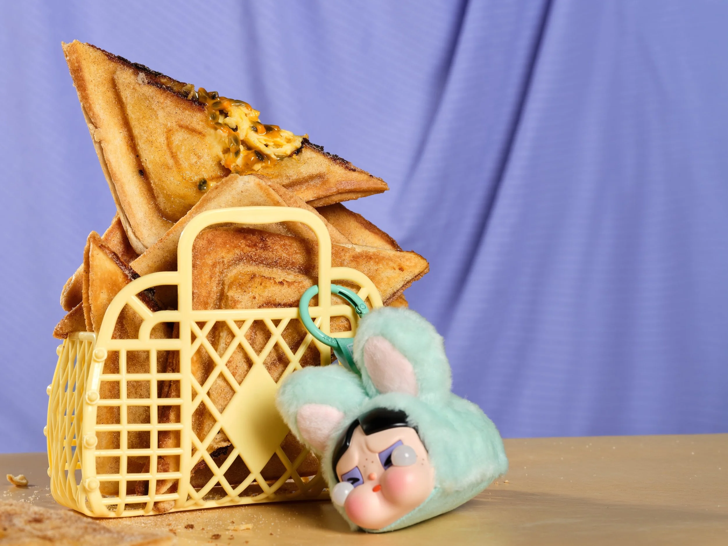 White chocolate, passionfruit and halloumi jaffles stacked in a small yellow basket, with a gooey passionfruit filling visible, set against a soft blue background with a pop mart crying again keyring beside them