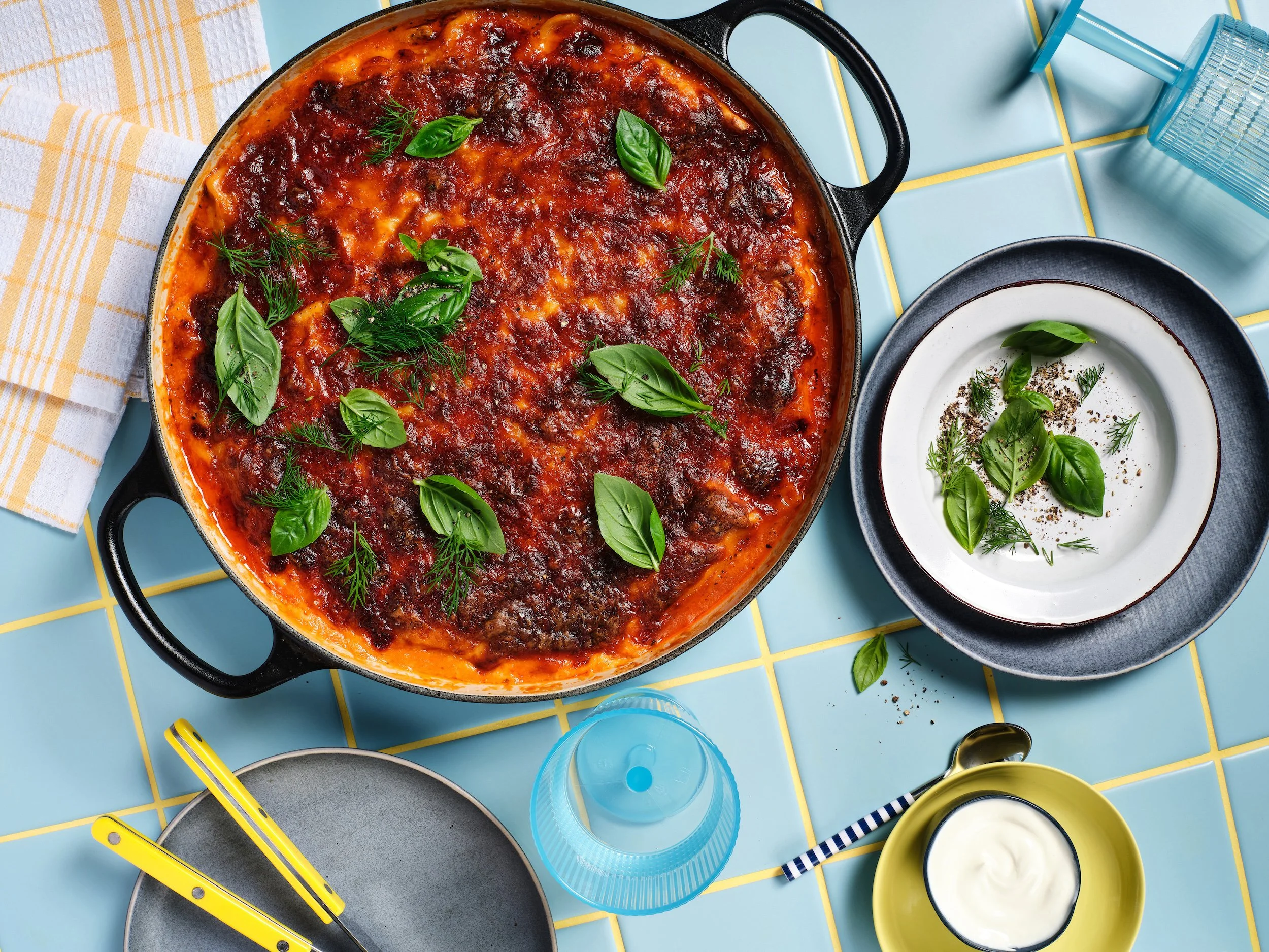 Pierogi lasagne baked in a black casserole dish with rich tomato sauce and fresh herbs, photographed overhead on a blue tiled table with serving plates.