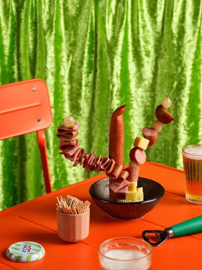 A sculptural arrangement of kabana, cheese cubes and cocktail onions skewered with toothpicks in a small bowl, styled like an ikebana display on an orange table with a beer glass, orange chair and lime green velvet backdrop.