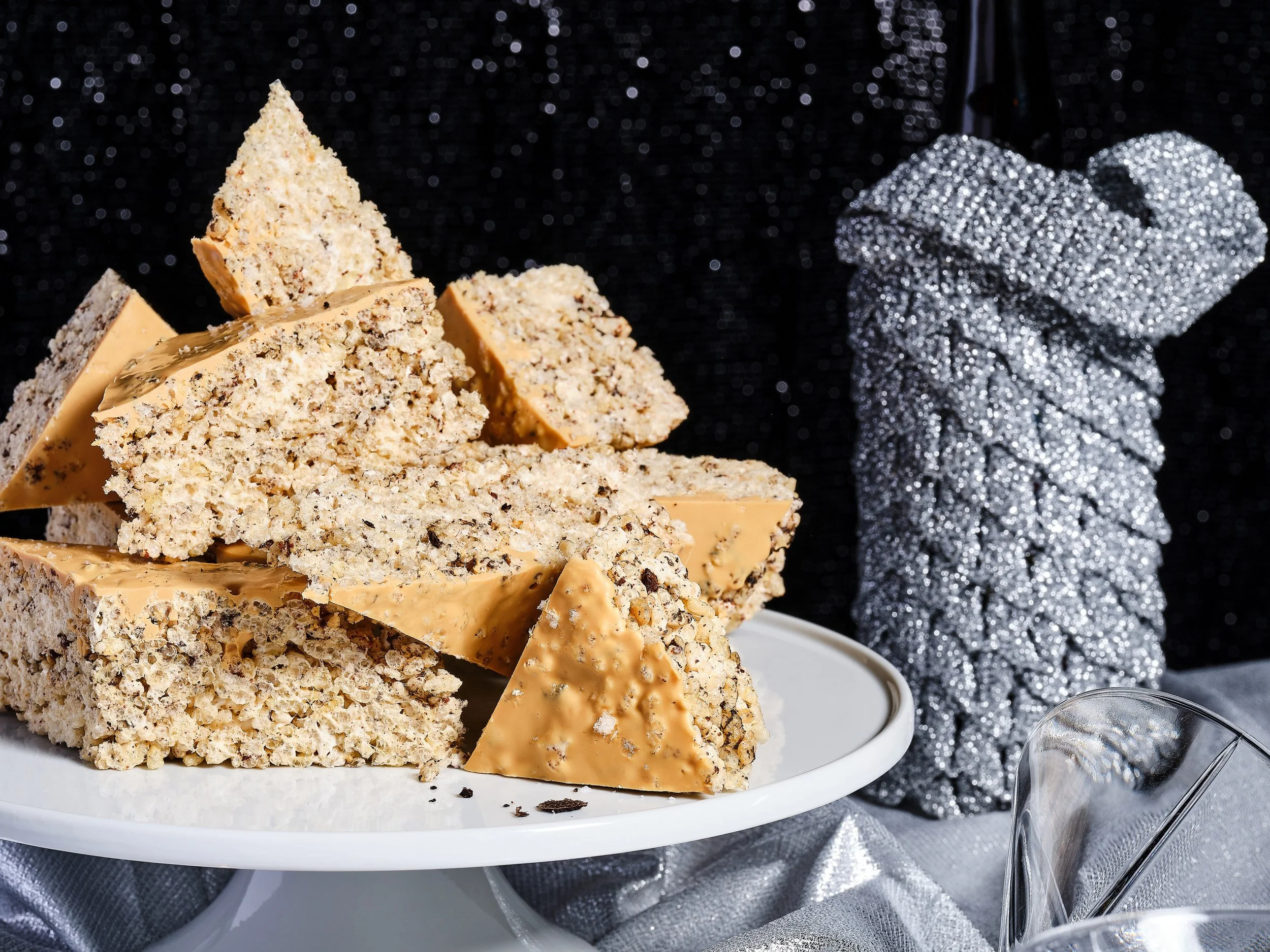 Stack of black truffle Rice Krispie treats coated in peanut butter Caramilk chocolate on a white cake stand, styled against a black sequin backdrop with silver draped fabric and crystal glassware
