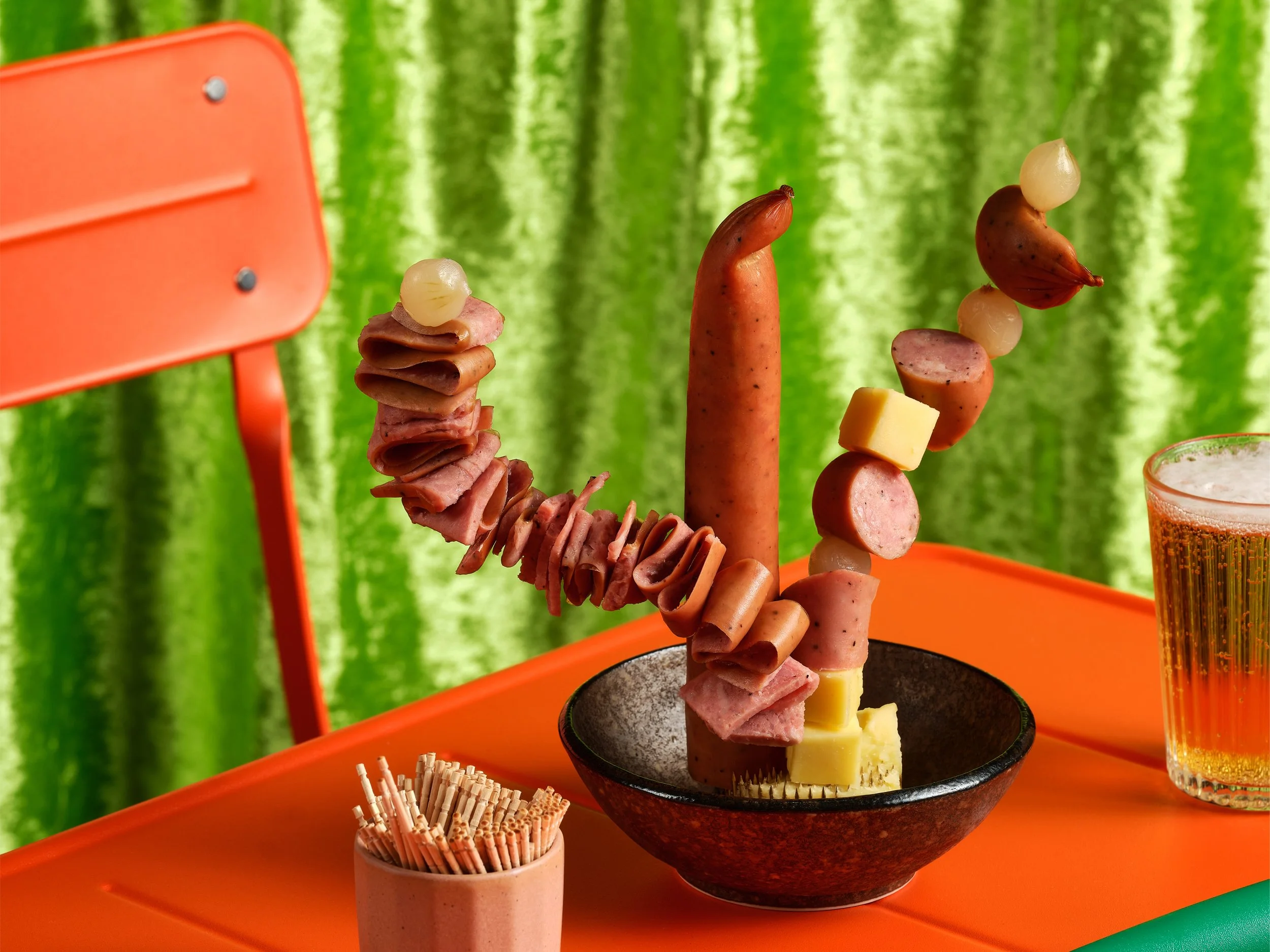 A sculptural arrangement of kabana, cheese cubes and cocktail onions skewered with toothpicks in a small bowl, styled like an ikebana display on an orange table with a beer glass, orange chair and lime green velvet backdrop.
