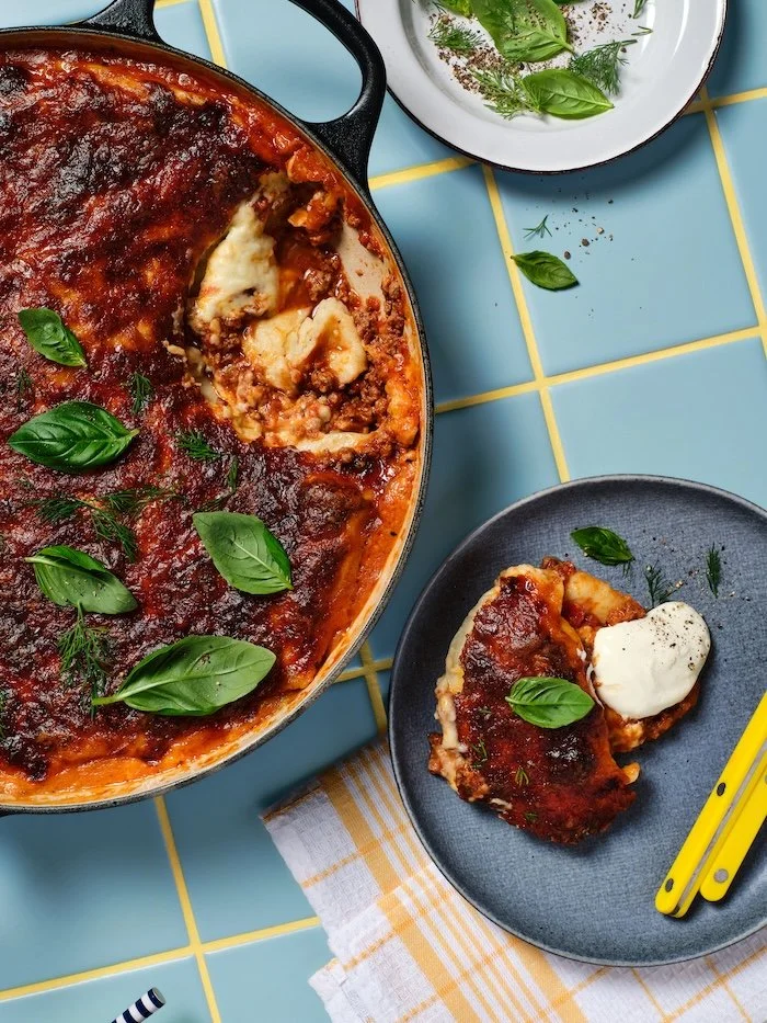 Overhead photo of pierogi lasagne in a black baking dish with a slice removed, topped with tomato sauce, basil and dill, with a plated serving on the side.