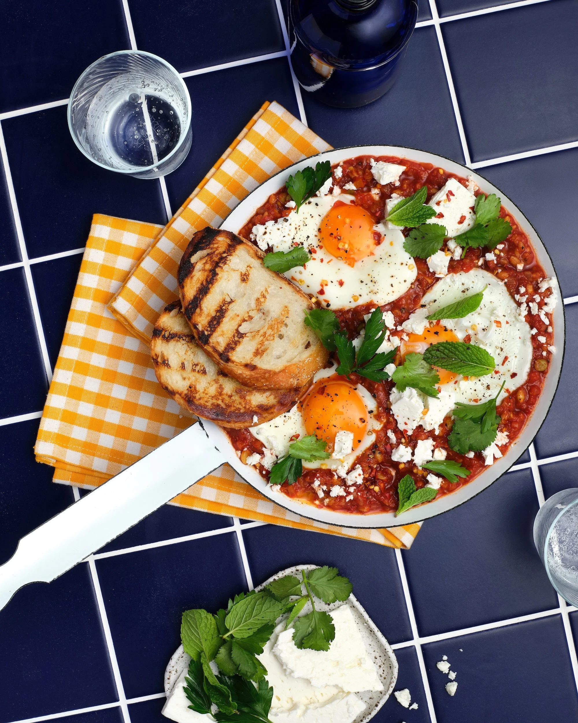 Overhead photograph of a skillet of shakshuka filled with rich tomato sauce and three sunny-side-up eggs, topped with crumbled feta, fresh parsley, coriander and mint. Thick slices of charred grilled bread rest across the pan, which sits on a bright 