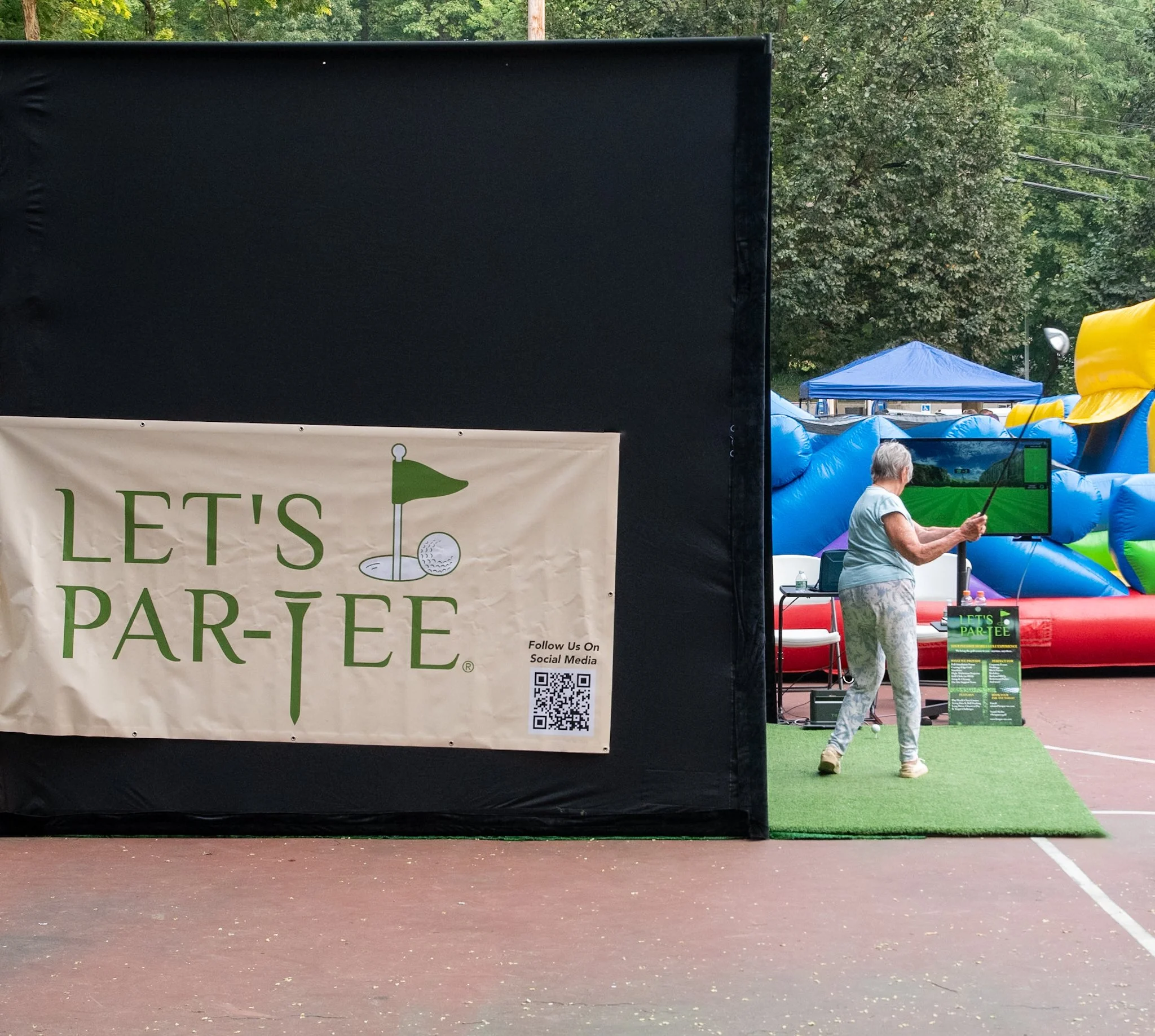 An outdoor event with a large inflatable slide in the background and a woman swinging a golf club in the middle. A black partition with a 'Let's Par-tee' sign and a QR code is in the foreground.