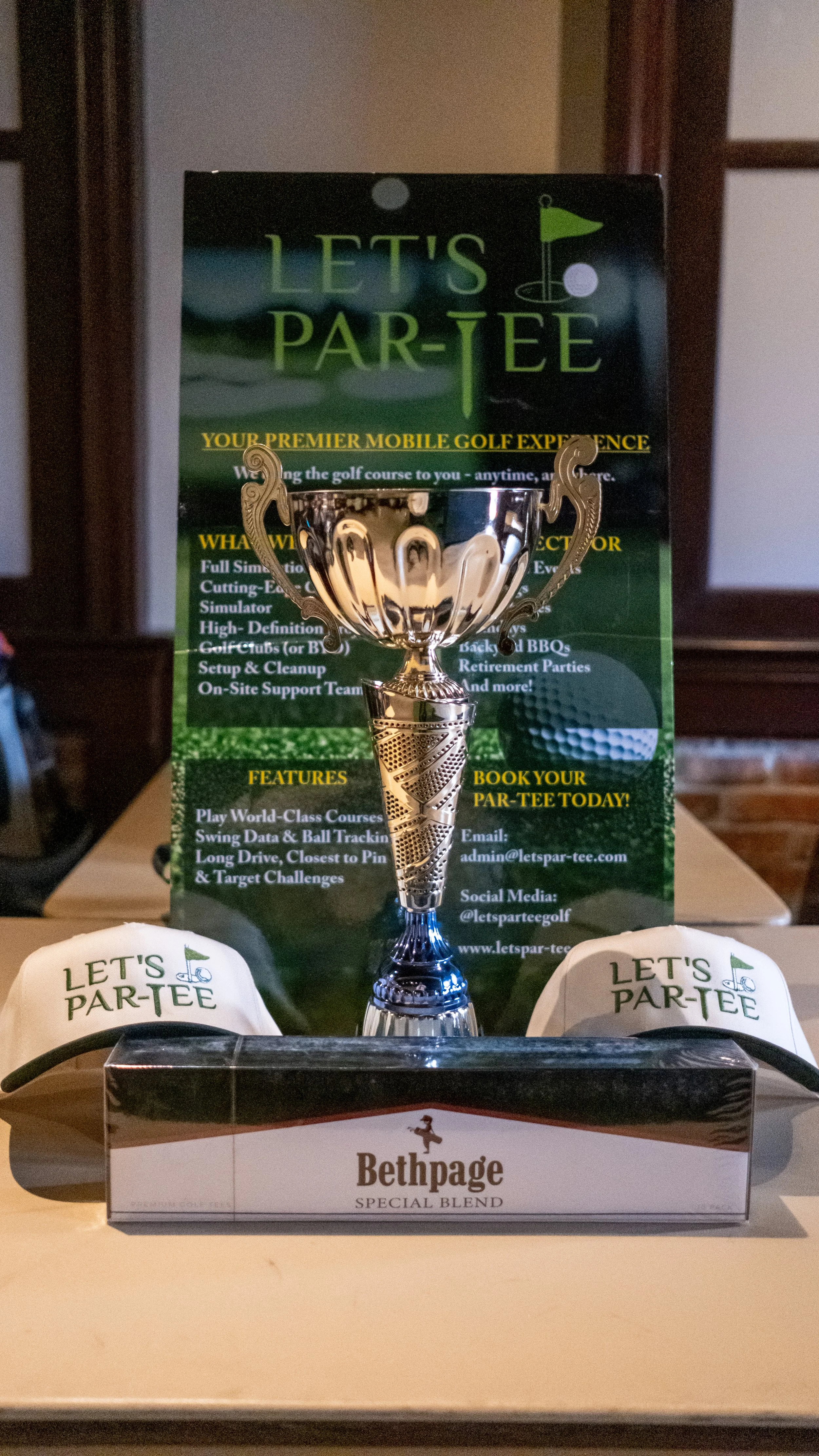 Golf trophy displayed on a table with promotional material behind it, white snap-back hats with 'Let's Par-Tee' logo on either side, and a box of Bethpage Special Blend golf tees in front.