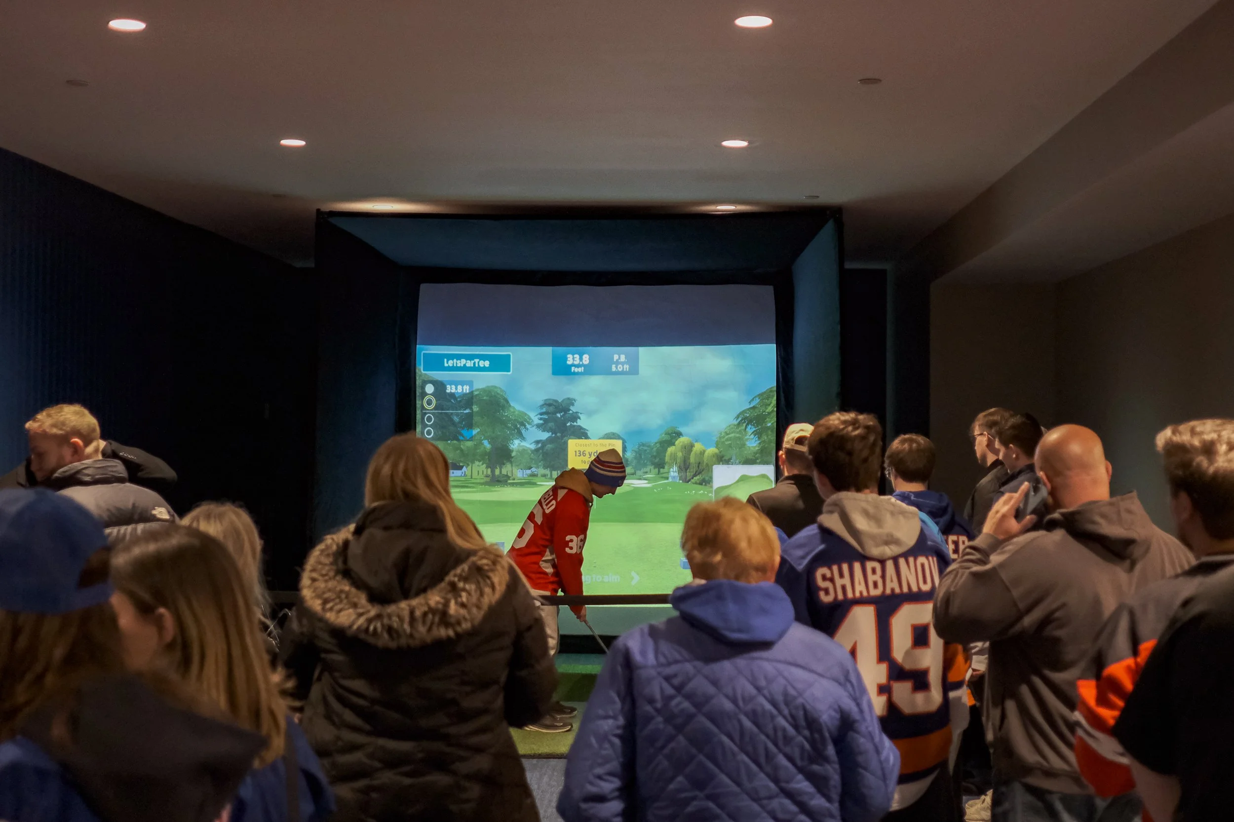 A group of people watching a golf simulation where a person is preparing to hit a ball in a virtual environment.