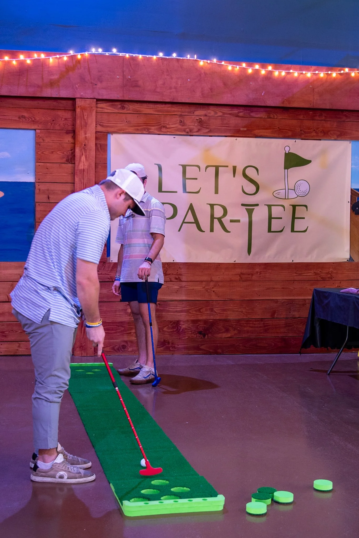 Two men playing mini golf on a green carpeted indoor course with a wooden wall and a sign that says "Let's Par-tee" in the background.