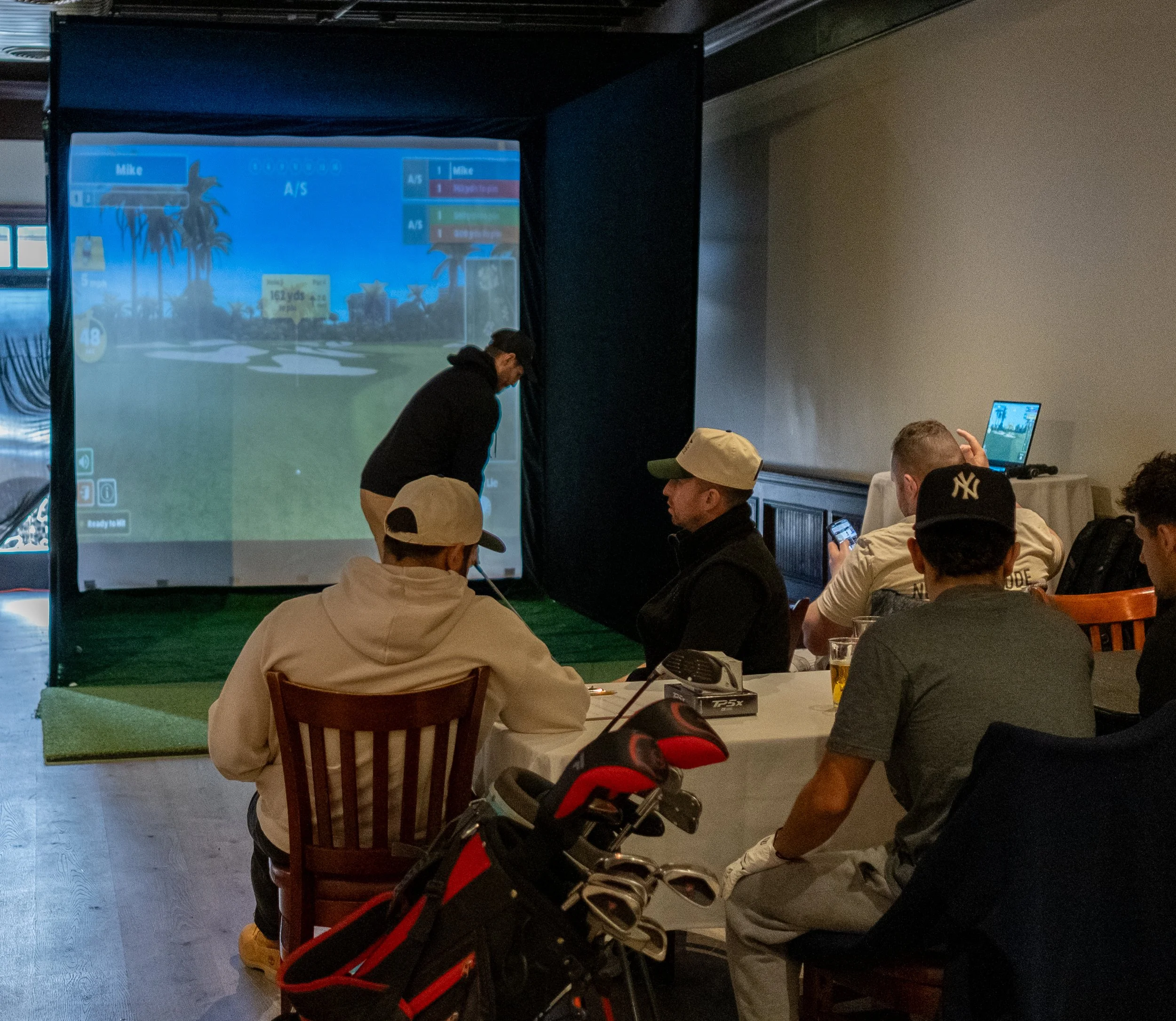 A group of people watching a golf simulator setup in a room, with one person actively playing on the screen and others sitting at a table, with golf clubs visible in the foreground.