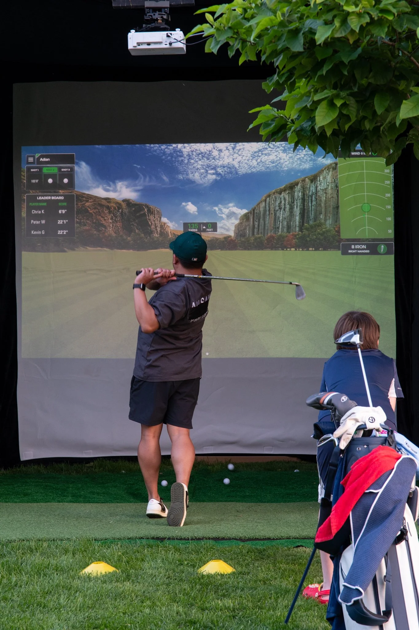 A person practicing golf using a digital golf simulator, with a virtual landscape on the screen and a golf bag with clubs nearby.